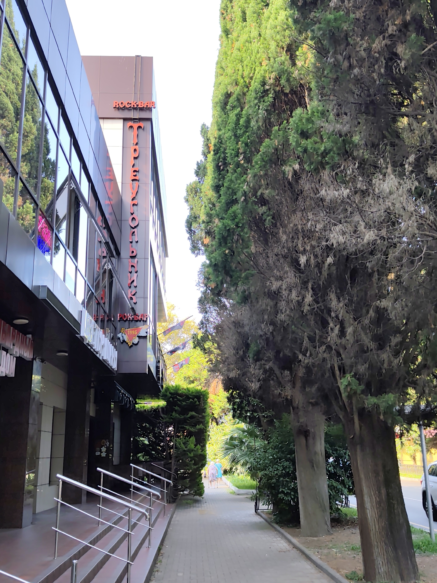 The image shows a modern building with glass windows and a sign that reads "ROCKBAR". The building is flanked by tall evergreen trees. A paved walkway runs alongside the building, bordered by a curb and trees. Two elderly women are walking on the walkway in the distance. The scene appears to be a street in Sochi, Russia, given the style of the building and the vegetation. The lighting suggests it is daytime, possibly late afternoon given the shadows.