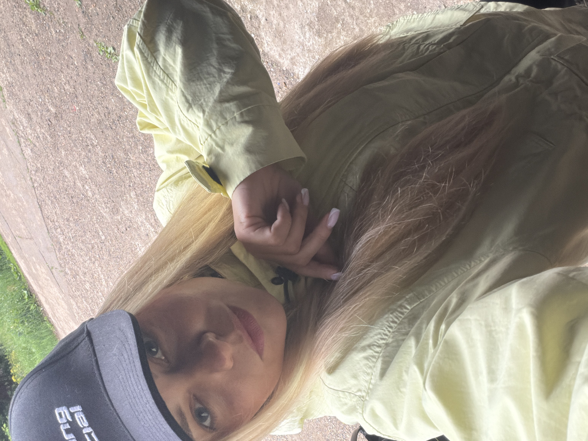 The scene, stated to be in Votkinsk, Russia, captures a close-up, high-angle selfie of a young adult female outdoors. She has long, straight blonde hair, which falls over her shoulders. She is wearing a black baseball cap with partial white lettering, including "ng" and "ficial," visible on the front. Her attire includes a light yellow or pale lime-green long-sleeved jacket with visible stitching and a black button detail on the cuff of her right arm. Her right hand, featuring light-colored nail polish on manicured nails, is gently posed near her chest with fingers slightly curled. She looks directly into the camera with a neutral expression. The background consists of a textured, light brown ground surface, possibly gravel or an unpaved path, with sparse green vegetation, and blurred green foliage in the upper left, indicating an outdoor setting under bright daylight.