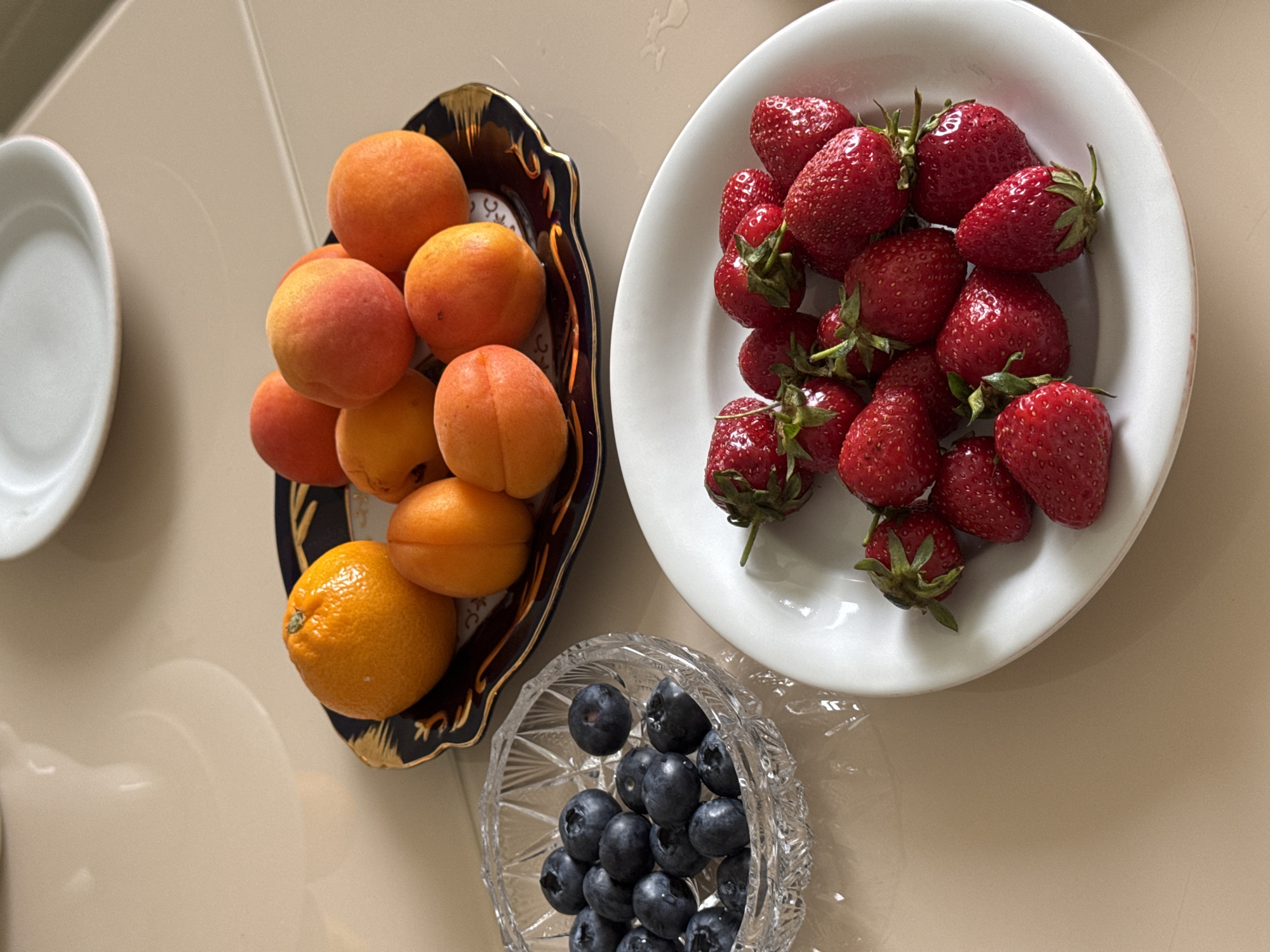 An overhead shot presents a vibrant still life of fresh fruits arranged on a light-colored, possibly tiled or polished, indoor surface, likely a kitchen or dining table in Izhevsk, Russia.
In the upper right, a white, slightly oval-shaped ceramic bowl holds a generous portion of ripe red strawberries. Each strawberry has its green calyx and stem intact, indicating freshness. The berries are plump and clustered together.
To the left of the strawberries, a dark blue decorative plate with an intricate gold rim and patterning displays a selection of orange fruits. These include approximately ten to twelve apricots, varying slightly in their orange and reddish blush, and a single, larger orange with a textured peel positioned towards the bottom-left of the plate. The plate itself has an ornate, wavy edge.
Below the decorative plate, a small, clear glass bowl with a cut-glass or crystal pattern contains a handful of dark blue blueberries. The berries are round and appear fresh.
Partially visible on the far left, a simple white ceramic plate or bowl contributes to the overall clean and bright aesthetic of the scene. The uniform lighting suggests a daytime setting, though specific time or weather cannot be determined. No people are visible, nor is any specific activity taking place beyond the arrangement of the fruits. There is no discernible text within the image. The composition highlights the natural colors and textures of the fruits, ready for consumption.