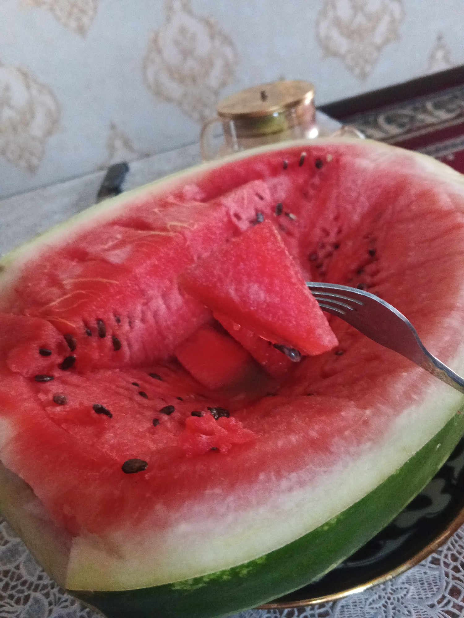 A close-up, horizontal shot shows a partially eaten watermelon with a fork piercing a slice. The watermelon is bright red, with visible black seeds scattered throughout the flesh and a white rind. The fork is made of silver metal and is positioned to the right of the frame, angled towards the watermelon. The watermelon rests on a dark plate with a decorative rim, which is on top of a white, lacy tablecloth. In the background, out of focus, a beige-patterned wallpaper is visible. The lighting suggests an indoor setting, possibly in Farkhad, Uzbekistan, with a hint of a metallic object, likely a teapot lid, in the upper right corner.