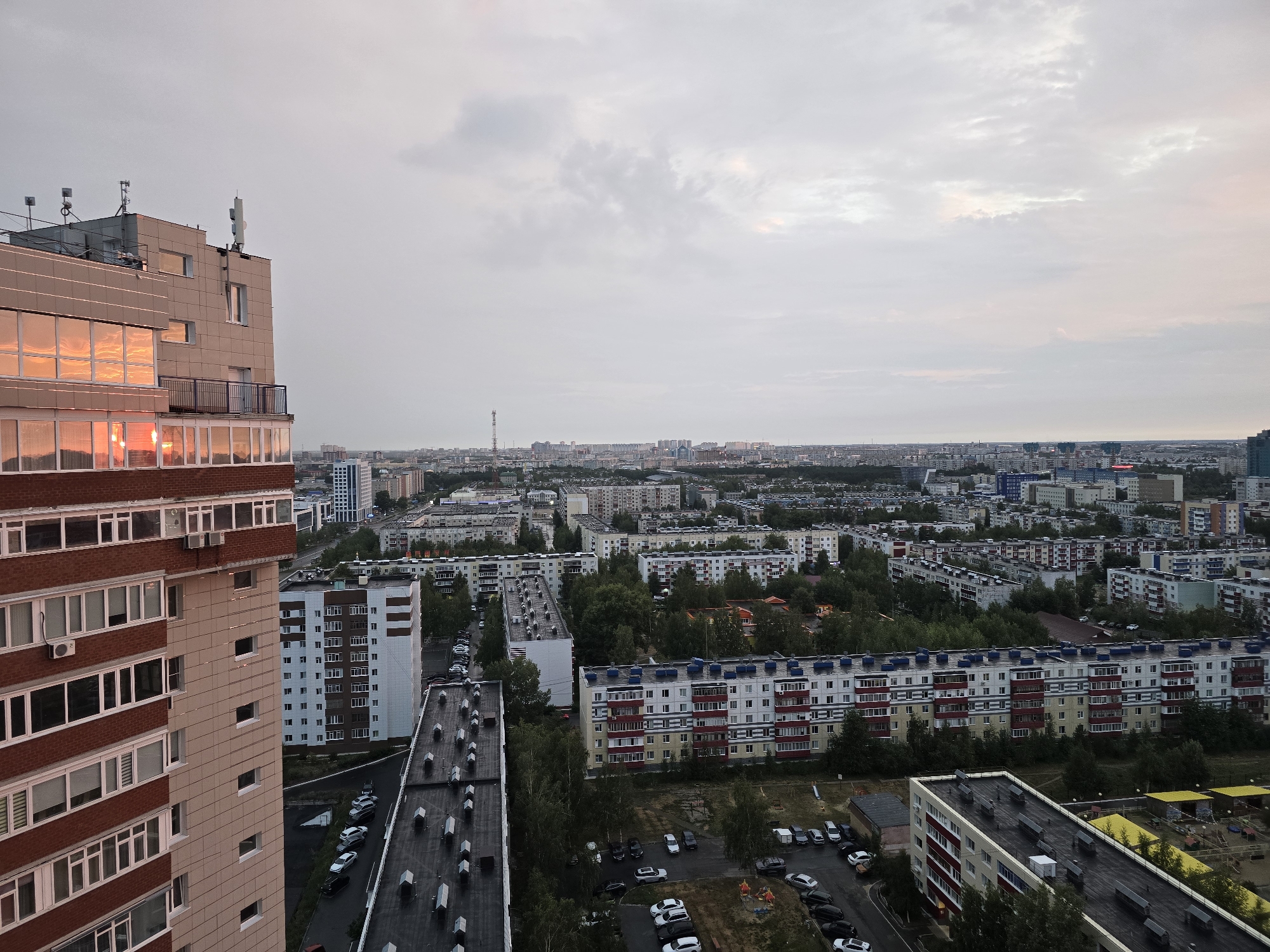 An aerial view shows a sprawling urban landscape at dusk in Surgut, Russia. The foreground is dominated by a tall, modern apartment building with a facade of beige panels and red brick, featuring numerous windows reflecting the warm hues of the setting sun. To the left of this building, a balcony is visible. Adjacent to the main building, other residential high-rises with distinctive white and red facades are lined up, interspersed with green trees and courtyards. Several cars are parked along the streets and in parking lots between the buildings. In the distance, the city stretches out, a dense collection of buildings under a cloudy sky. The overall impression is of a residential district in a vast Russian city.