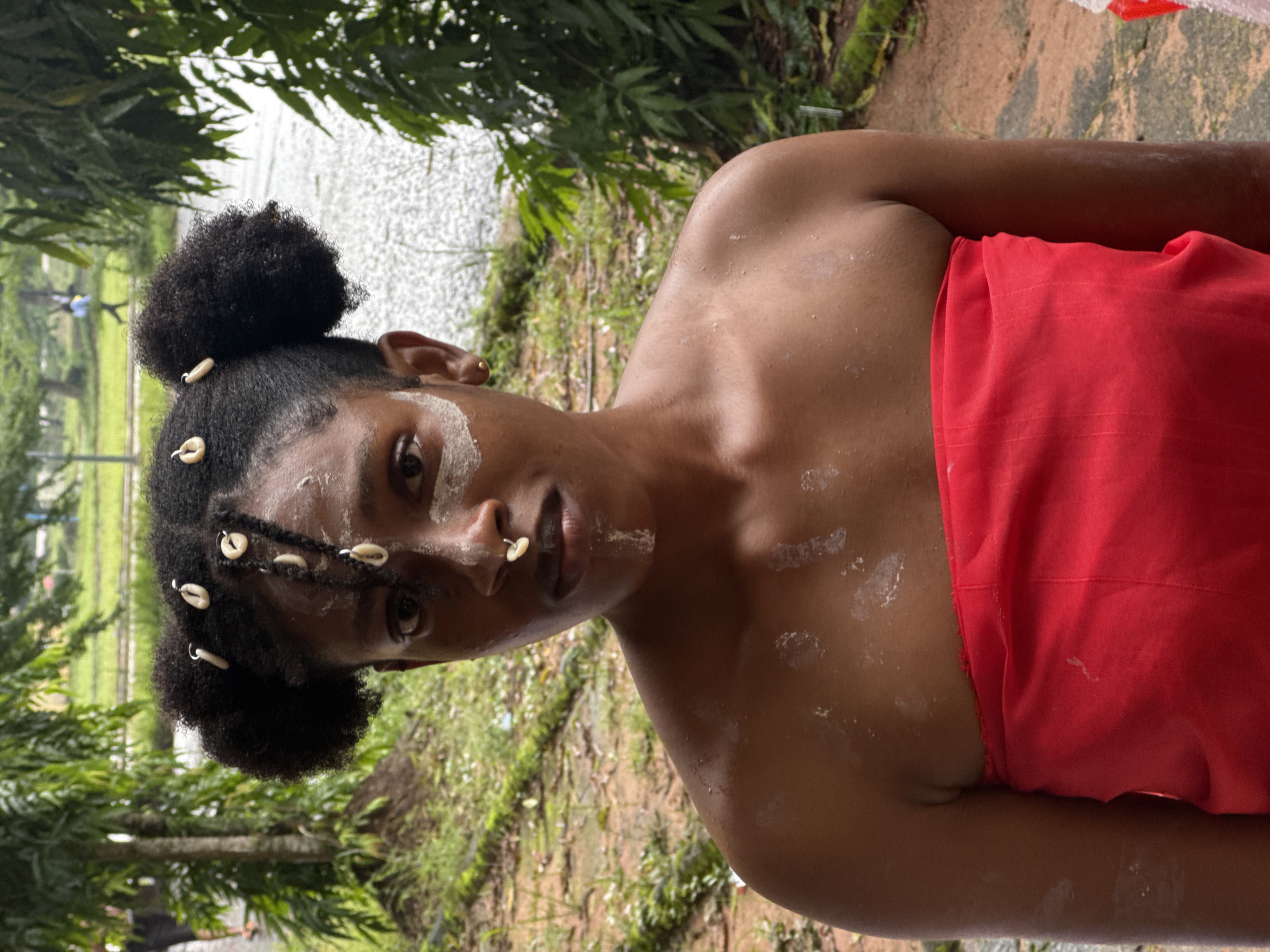 A young woman with dark skin, wearing a red cloth around her chest, poses for a portrait. She has her hair styled in two large puffs, adorned with cowrie shells and white beads. White paint is applied to her face, with streaks on her cheeks and forehead, and a shell is placed on her nose. The background shows an outdoor environment with trees, foliage, and what appears to be a paved or muddy path. The overall lighting suggests an overcast day. The location is identified as Ihiagwa, Nigeria.