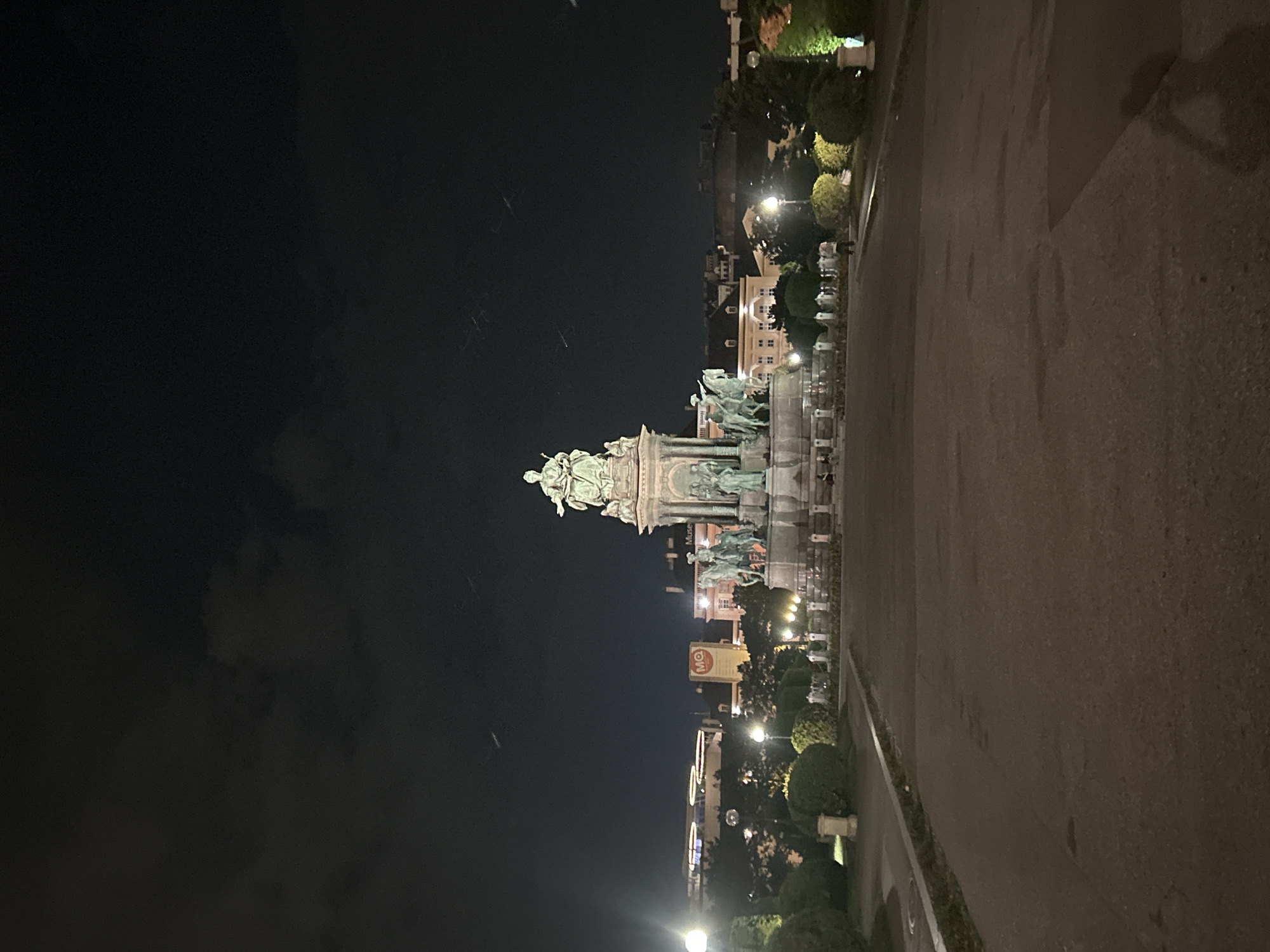 The image depicts the Maria-Theresien-Platz in Vienna, Austria at night. The central focus is the monument of Empress Maria Theresa, a grand equestrian statue, illuminated and surrounded by sculptures of allegorical figures. In the background, several buildings are visible, including the Museum of Natural History and the Museum of Art History, their architectural details softened by the darkness. The sky above is dark with some scattered clouds and faint streaks, possibly from aircraft. The foreground shows a wide paved area, leading towards the monument, with manicured bushes and lampposts lining the pathway. A few individuals are sparsely present, some sitting near the monument and one walking in the middle distance. The overall atmosphere is one of nocturnal grandeur and historical significance.