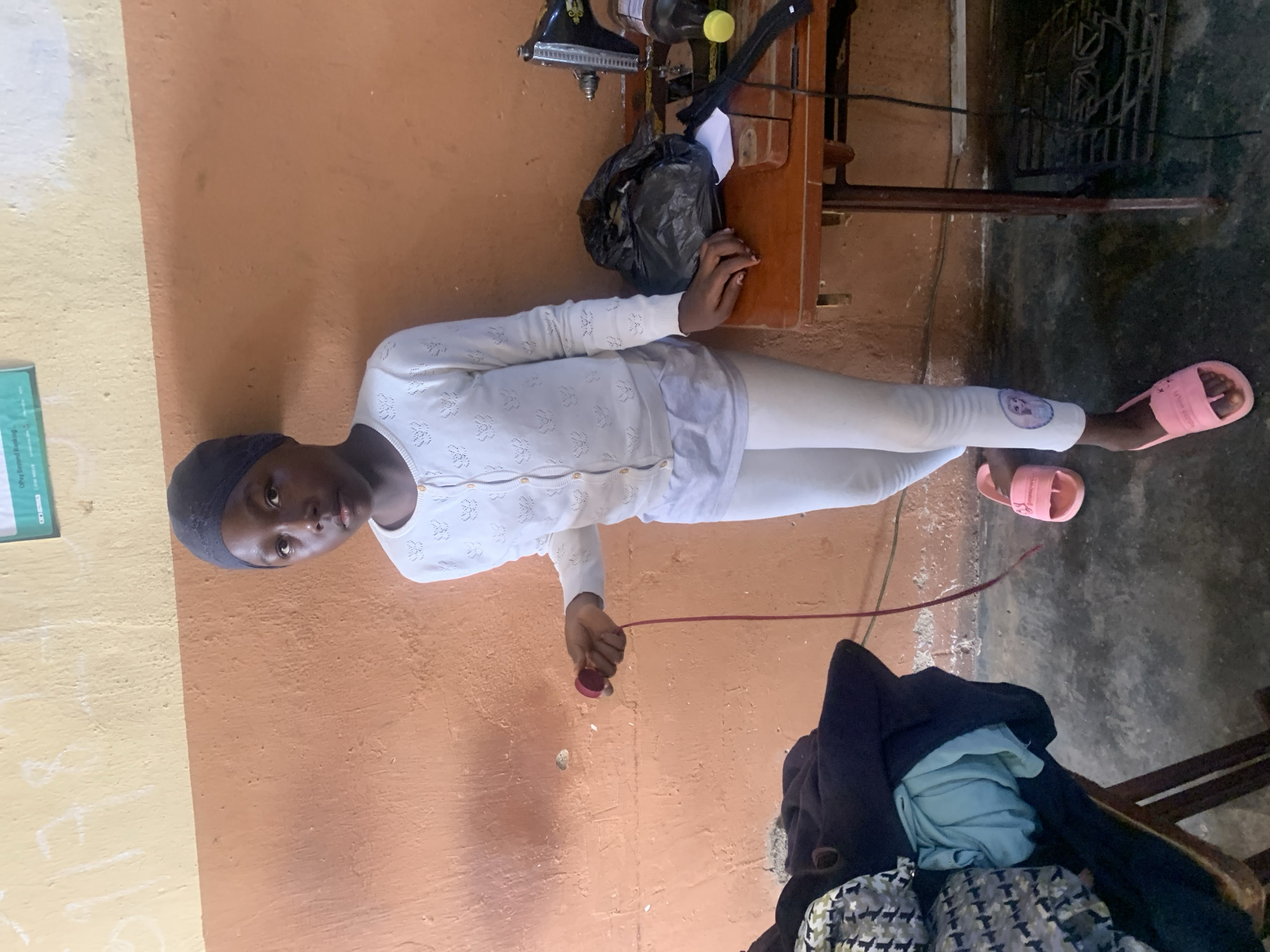 A young Black girl wearing a dark blue headscarf, a white cardigan with a patterned print, and white leggings with a graphic on the left leg stands in front of a textured, peachy-orange wall. She is holding a red cord with a circular object attached to its end. To her right, a wooden table holds a black sewing machine, a black trash bag, a bottle of yellow liquid, and a black zipper. Next to the table on the left, a wooden chair is covered with folded dark blue and patterned fabric. The floor is a rough, gray concrete. The overall setting appears to be a simple, interior workspace.
