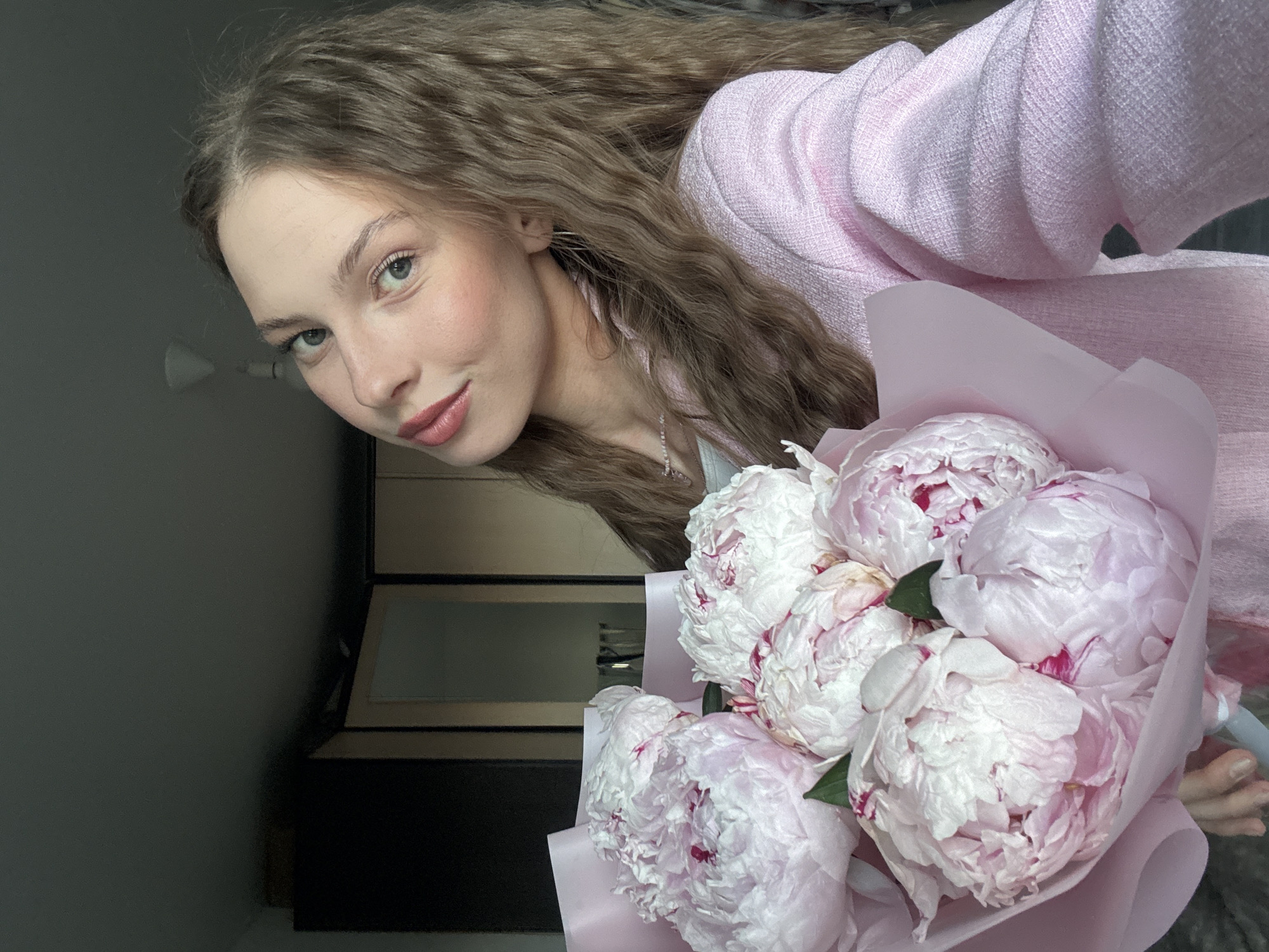 A young woman with long, wavy brown hair and light blue eyes smiles at the camera. She is wearing a soft pink textured jacket and a delicate beaded necklace. In her right hand, she holds a bouquet of pale pink peonies wrapped in pink paper. The background appears to be indoors, with a dark gray wall on the left and a dark wooden cabinet with reflective surfaces on the right. The lighting suggests it is daytime, possibly late morning or early afternoon, with soft, diffused light. The woman's expression is friendly and gentle, and the overall scene evokes a feeling of warmth and perhaps a special occasion, given the presence of the flowers. There is no visible text in the image.