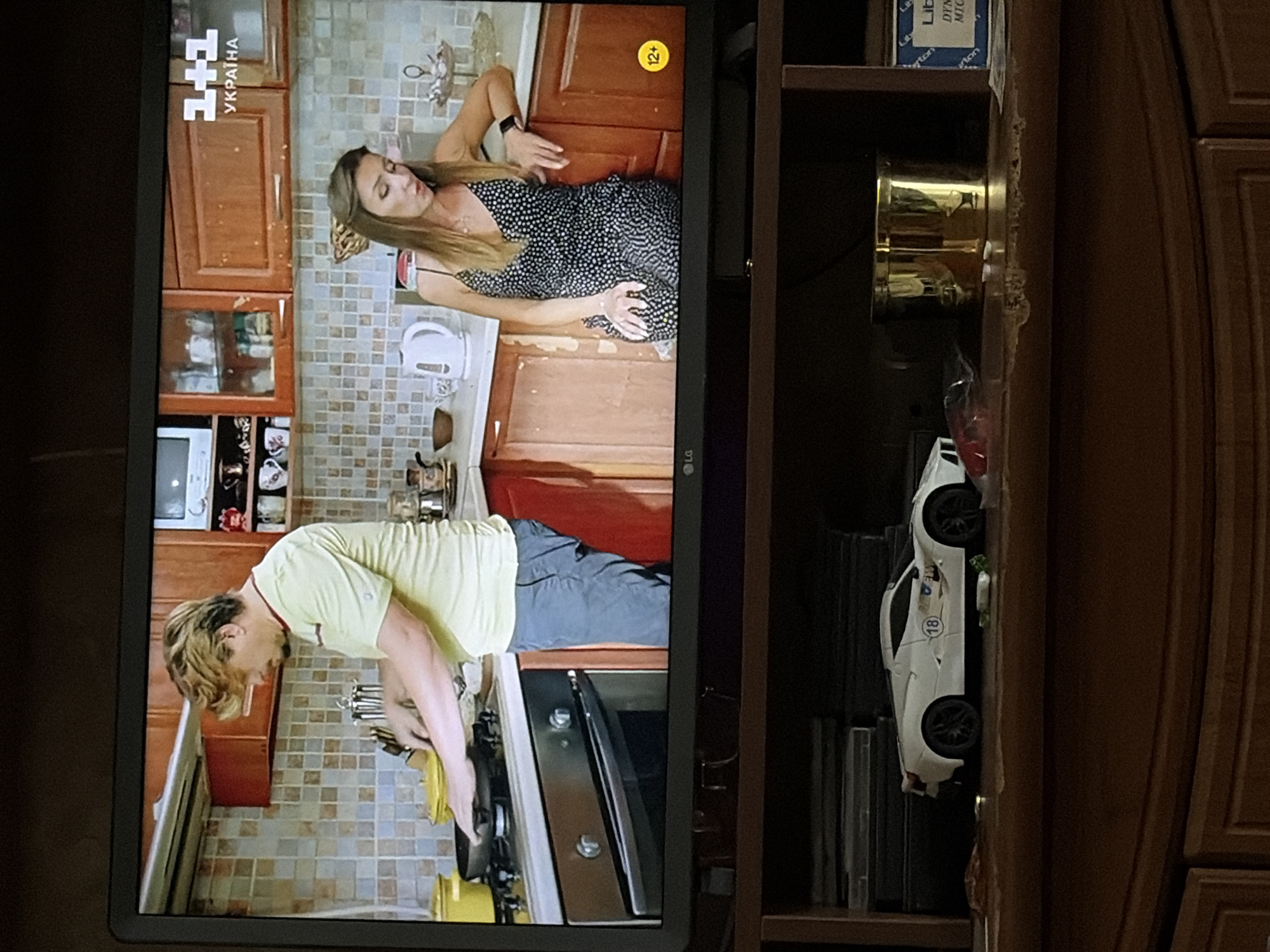The image shows a television screen displaying a cooking show, with a man and a woman in a kitchen setting. The man, with blond, wavy hair, is wearing a yellow t-shirt and blue jeans, and is standing at a stove, seemingly cooking. The woman, with long brown hair, is wearing a black and white polka dot dress and is seated at a counter, looking towards the man. In the foreground, below the television, is a wooden cabinet with a white toy sports car on its shelf, along with a golden cup and some other items. The scene is indoors, likely a living room or entertainment area. The specific location of the city is unknown.