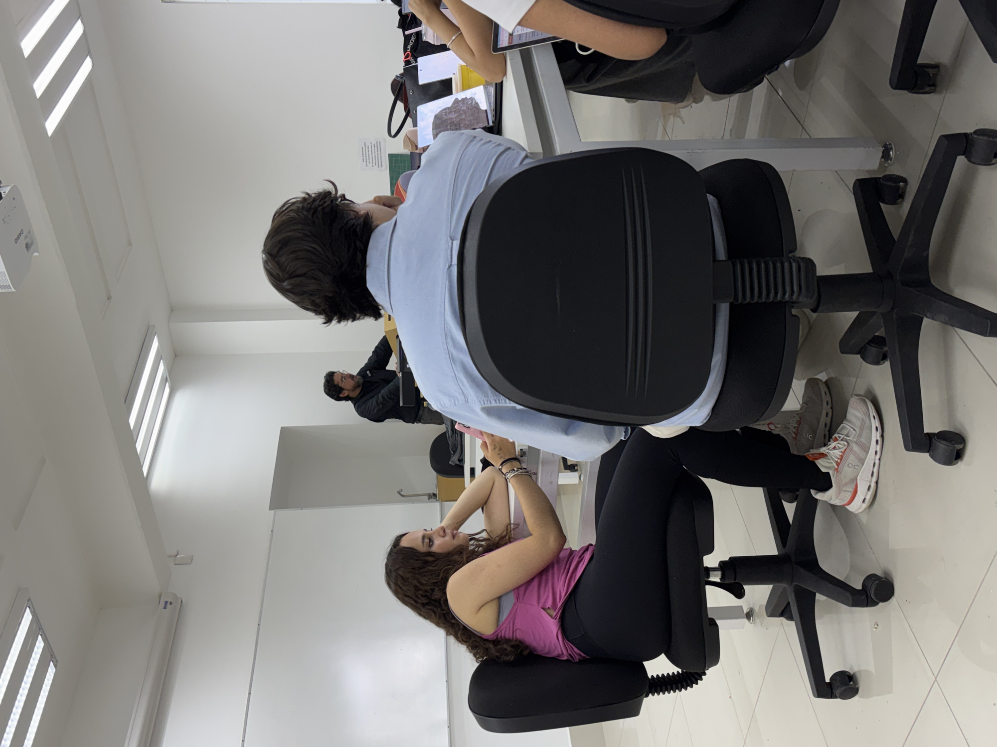 The image shows a classroom scene in San Andrés Cholula, Mexico. Four people are visible: a woman with curly hair sitting on a black office chair, wearing a pink tank top and black leggings, is looking at a phone. A man with dark hair is seated in a black office chair, wearing a light blue shirt. Another person seated at the table is using a tablet. There is also a person walking in the hallway with glasses, wearing a dark jacket. The classroom has white walls and a white tiled floor. The desks are white with metal frames. There are overhead fluorescent lights. A projector is mounted on the ceiling.