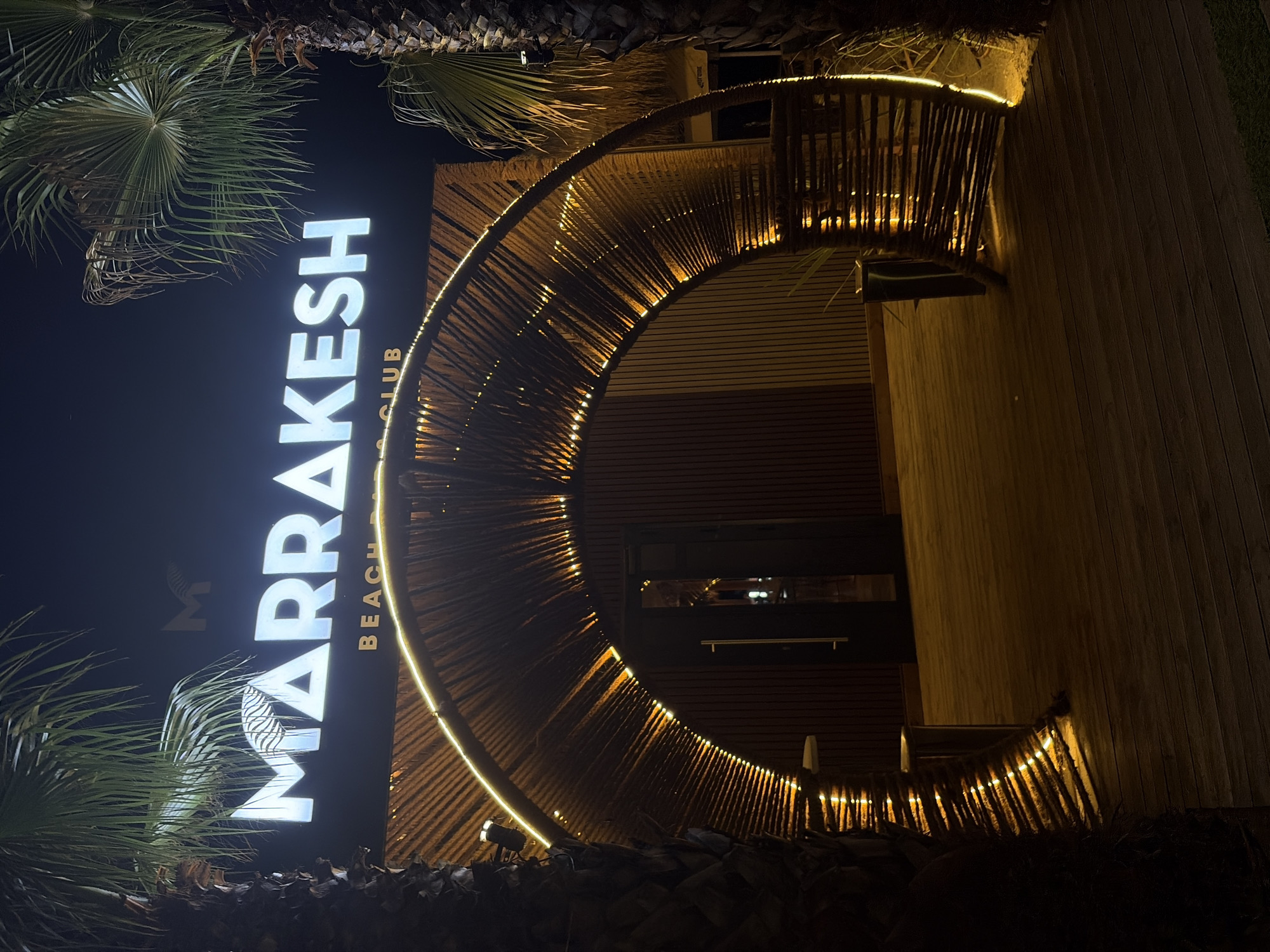 This is a night-time exterior shot of a beach club called "MARRAKESH BEACH CLUB" located in Burgas, Bulgaria. The prominent subject is the illuminated sign for the club, with large, white, block letters spelling out "MARRAKESH". Below it, in smaller, gold-colored text, are the words "BEACH CLUB".
The building features a distinctive entrance with a curved, overhead structure constructed from vertical wooden poles, accented with a string of warm-toned fairy lights that create a glowing effect. This structure appears to be a decorative canopy or entryway. The building itself has a modern facade with horizontal wooden paneling.
Palm trees are visible in the foreground and framing the left side of the image, their fronds silhouetted against the dark night sky. The ground in front of the entrance is a wooden deck. The lighting suggests it is evening or night, and there is no indication of weather conditions. The overall atmosphere conveyed is one of a vibrant, possibly lively, night-time venue. There are no people visible in the image.