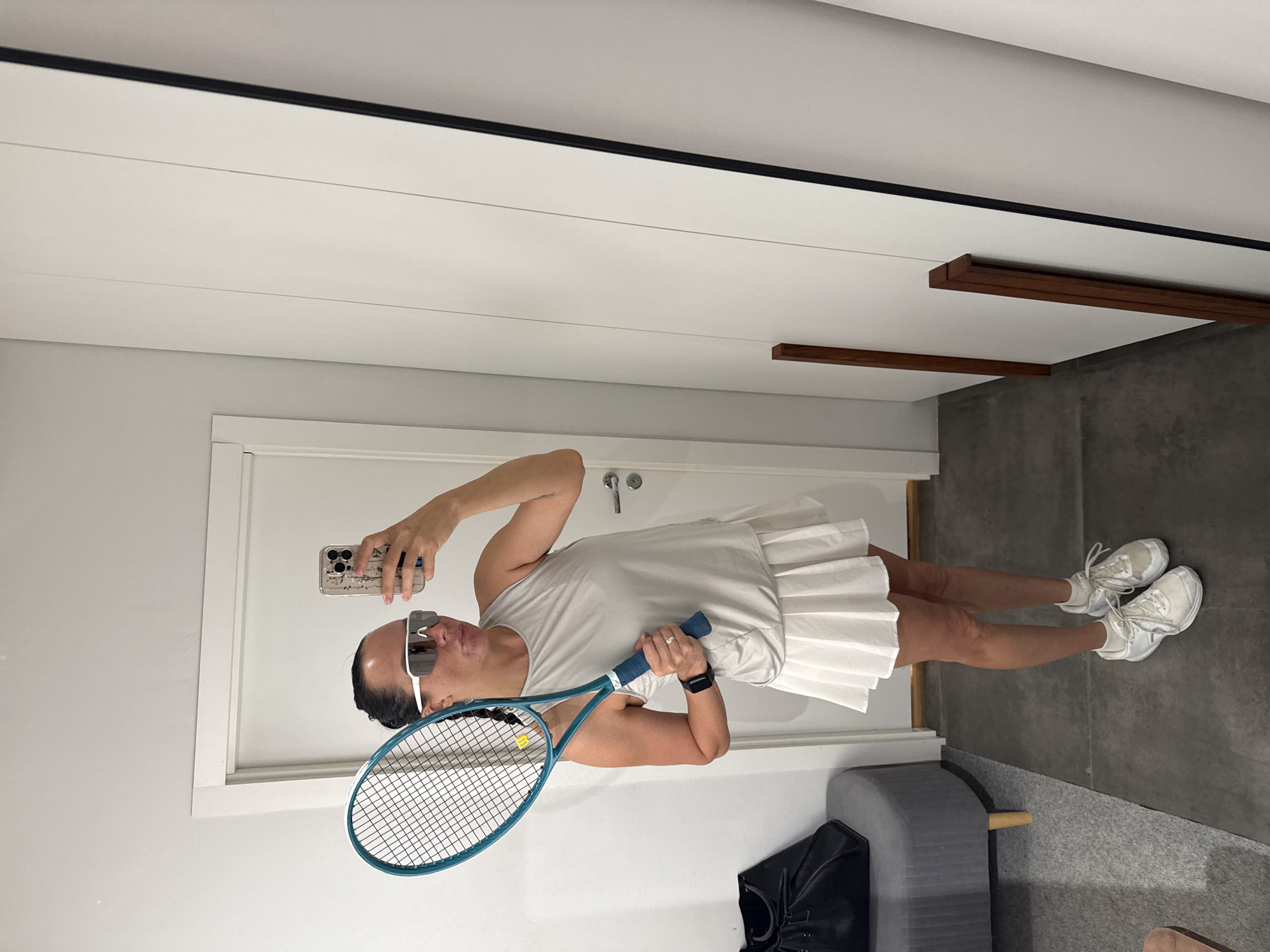 A woman stands in what appears to be a room with a white door and a large white mirrored closet. She is holding a tennis racket with a teal frame and blue grip. She is wearing white athletic shoes, white socks, and a light gray sleeveless top with a white pleated skirt. She has on large white wraparound sunglasses. She is taking a selfie with her phone, which has a floral case. To her left, there is a dark gray upholstered ottoman with wooden legs, and a black bag on top of it. The floor is a dark gray tile.