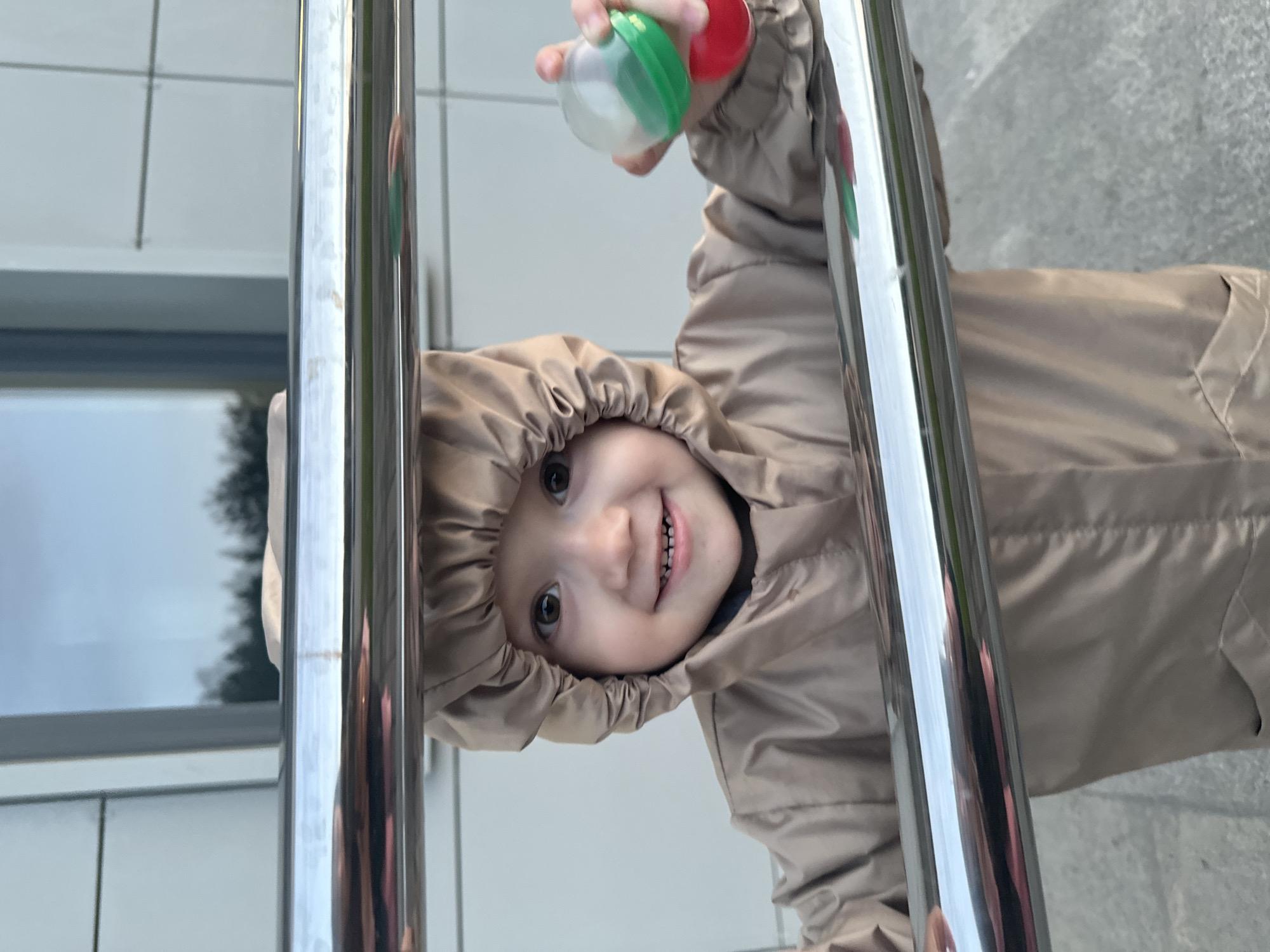 A young child, appearing to be around 2-3 years old, is positioned between two horizontal chrome railings, looking directly at the camera. The child is wearing a tan hooded jacket. Their right hand holds a clear plastic capsule toy with green and red components visible. The background consists of a modern building facade with a large window reflecting trees and the sky. The ground beneath appears to be paved. This scene was captured in Yekaterinburg, Russia.