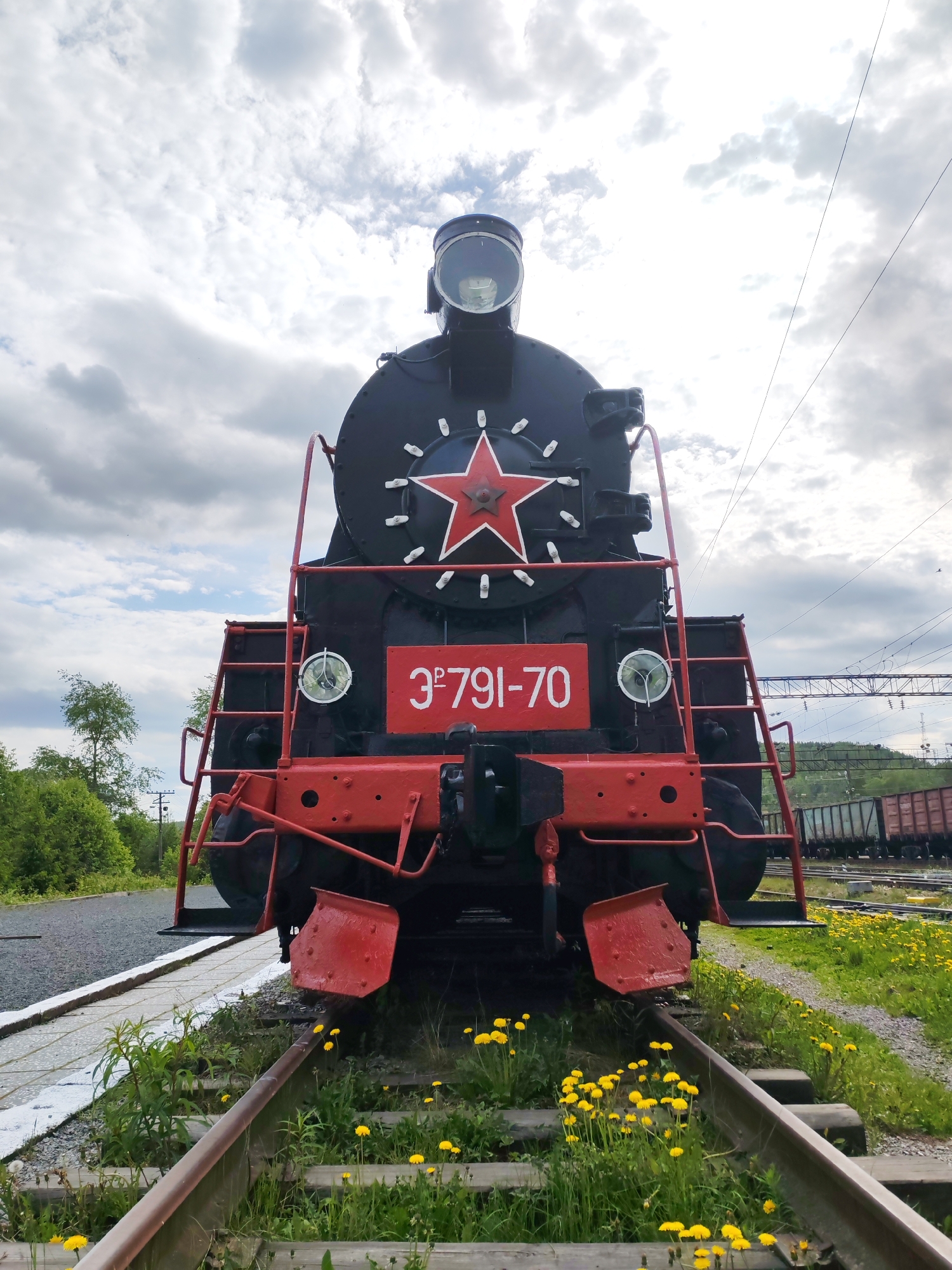 The image captures a head-on, low-angle view of a black steam locomotive. Its front features a large red five-pointed star with a white outline, centered on the boiler door, just below a prominent, circular headlamp. Below the star, a red rectangular plate displays the identification number "3ᵉ 791-70" in white Cyrillic characters and numerals. Two smaller, circular headlights are positioned on either side of this number plate. The locomotive's lower section incorporates red buffer beams and sturdy red-painted metalwork, including steps and handrails.
The locomotive is situated on a single track, which is partially overgrown with vibrant green grass and numerous bright yellow dandelions in the immediate foreground, suggesting a peaceful, perhaps disused, railway setting. To the left, a concrete platform edge runs alongside the track, leading to a gravel path. In the background, more train tracks are visible, along with several dark freight cars, indicating a railyard or station area. Distant green trees and foliage line the horizon, and utility poles with overhead power lines can be faintly seen against the sky. The sky is predominantly cloudy, with significant white cloud cover suggesting an overcast daytime. No people are visible in the scene. The overall impression is one of a historical piece of machinery set against a natural, slightly wild backdrop. Given the location context, this is likely a locomotive from the Russian railway system.