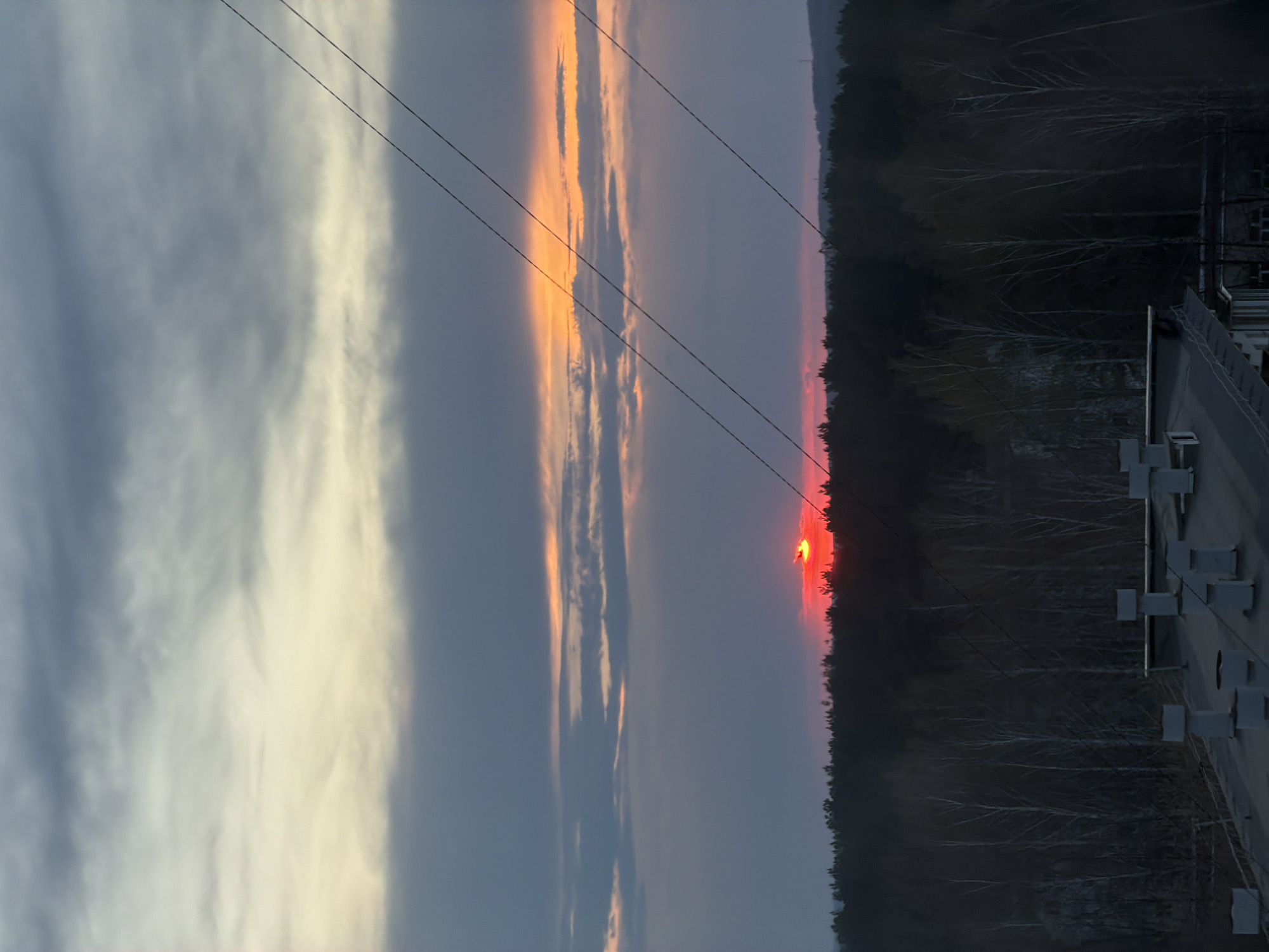 The image shows a sunset scene in the city of Chaikovsky, Russia. The sun is setting behind a dark tree line, creating a vibrant orange and red glow on the horizon. Above the horizon, the sky is a mix of blue and orange hues, with streaks of white clouds illuminated by the setting sun. Power lines stretch across the sky from left to right. On the right side of the image, there is a structure with multiple white rectangular objects, possibly a building or part of a dam. The overall environment appears to be a blend of natural and urban elements, with the forest meeting the man-made structure in the foreground.