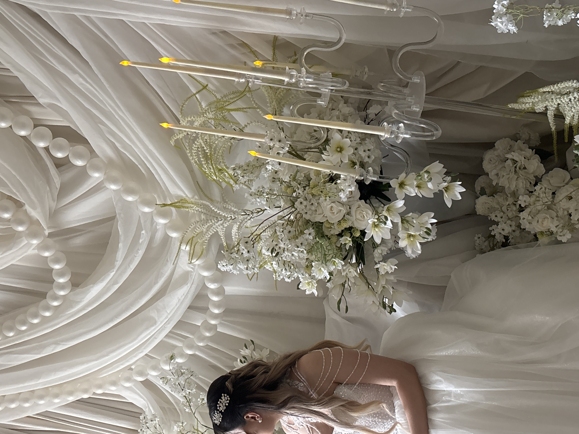 The image shows a woman lying on a white surface, possibly a bed or draped fabric. She is wearing a white dress with beaded straps and a dark hair accessory. Above her, there is an elaborate floral arrangement of white flowers and greenery, along with decorative strands of white pearls. Simulated candles on clear holders are interspersed within the flowers and fabric, providing illumination. It could be part of a wedding or formal event. The setting does not contain any information that is specific to the city of Yerevan, Armenia.