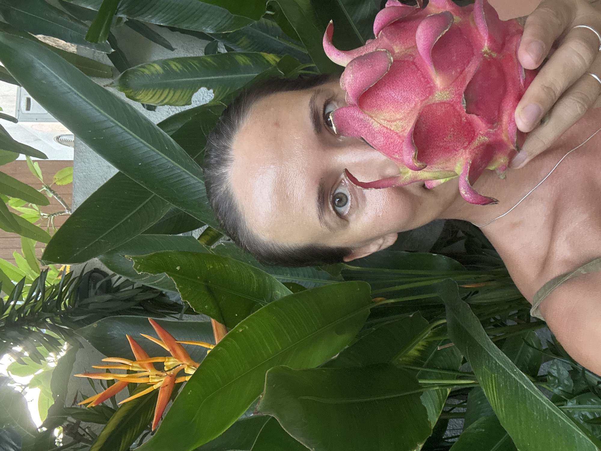 A person is holding a dragon fruit up to their face, standing among lush green foliage. The scene includes a variety of plants, including banana leaves and an orange flower. The location is Kuta Selatan, Indonesia. The person appears to be posing playfully with the fruit.
