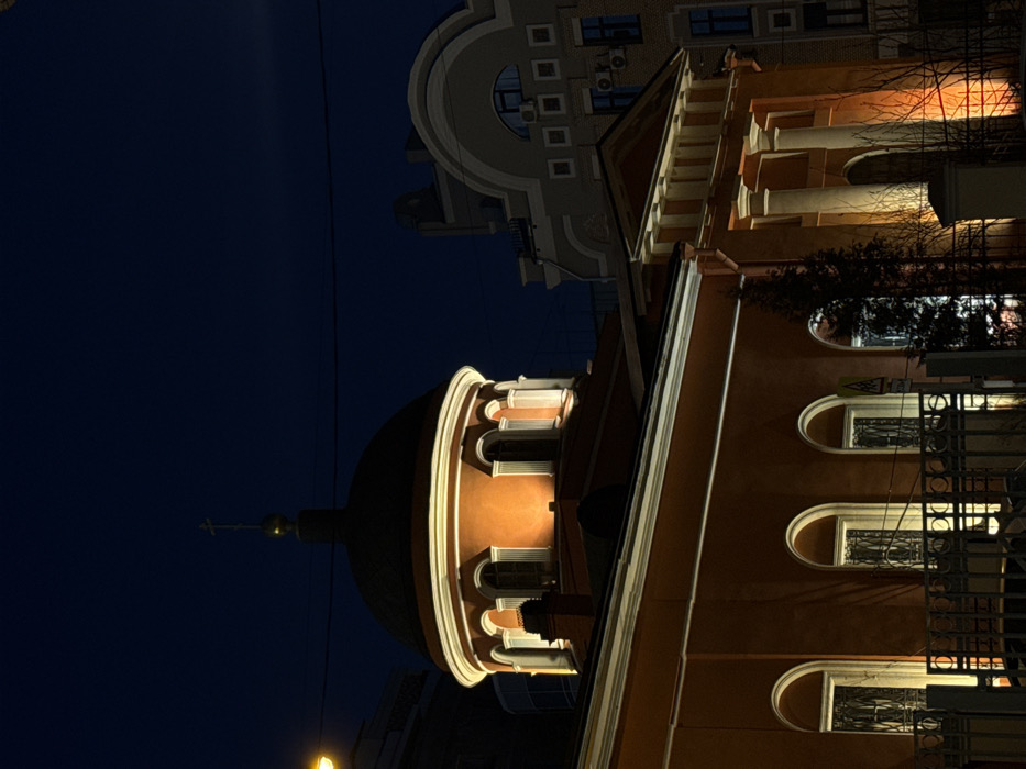 The scene captures the Church of the Exaltation of the Holy Cross, located at Vozdvizhenka Street 16, Moscow, Russia, during nighttime. The church presents an orange-red facade with prominent white architectural detailing, including cornices, arched window frames, and a portico on the right featuring two white columns and a triangular pediment. A large, dark dome, capped with a golden finial and an Orthodox cross, surmounts the structure. The drum beneath the dome displays four arched windows, all illuminated from below. Artificial lighting strongly illuminates the church's exterior, producing an orange-yellow glow on portions of the facade and columns. A dark metal fence runs across the lower foreground, partially obscuring the base of the church. A triangular yellow road sign with a red border is discernible behind the fence. To the immediate right of the church stands a taller, lighter-colored building with a distinct rounded facade at its top, featuring multiple square windows and visible external air conditioning units. Sparse, bare vegetation is present in the mid-ground, and a streetlamp is visible on the far left. The sky is dark blue. No individuals or discernible activities are present within the frame.