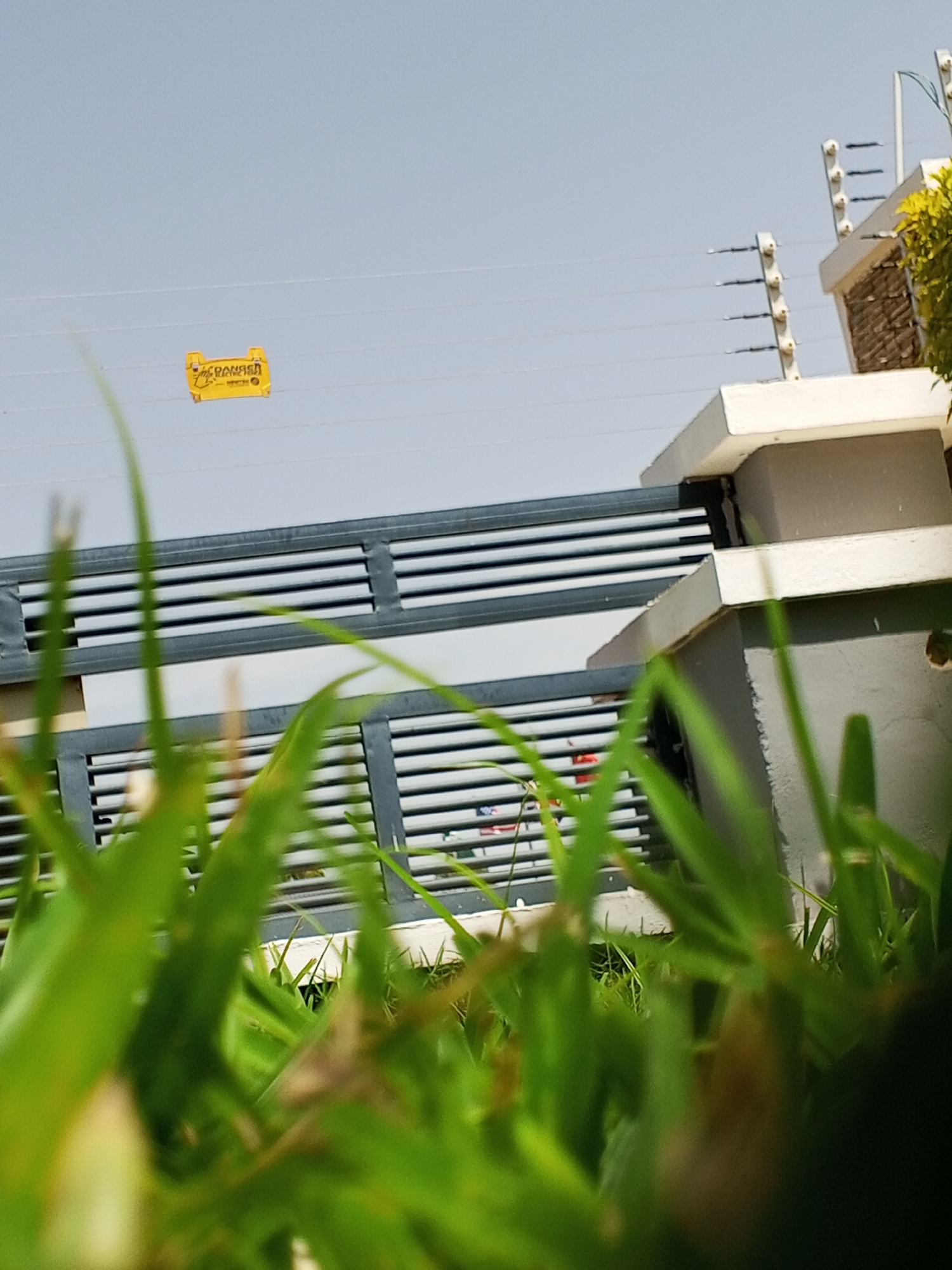 In a scene dominated by green foliage in the foreground, a partially obscured view of a gate and an electric fence is presented against a clear sky. The gate, made of dark metal slats, is visible behind the grass. Above the gate, a bright yellow sign with the word "DANGER" in black lettering is attached to what appears to be a wire or cable. Further in the background, the top of a building is visible, featuring a white ledge and brickwork, with a section of an electric fence mounted on poles. The scene is set outdoors with no people or specific actions captured. The location is identified as Unknown, Unknown.