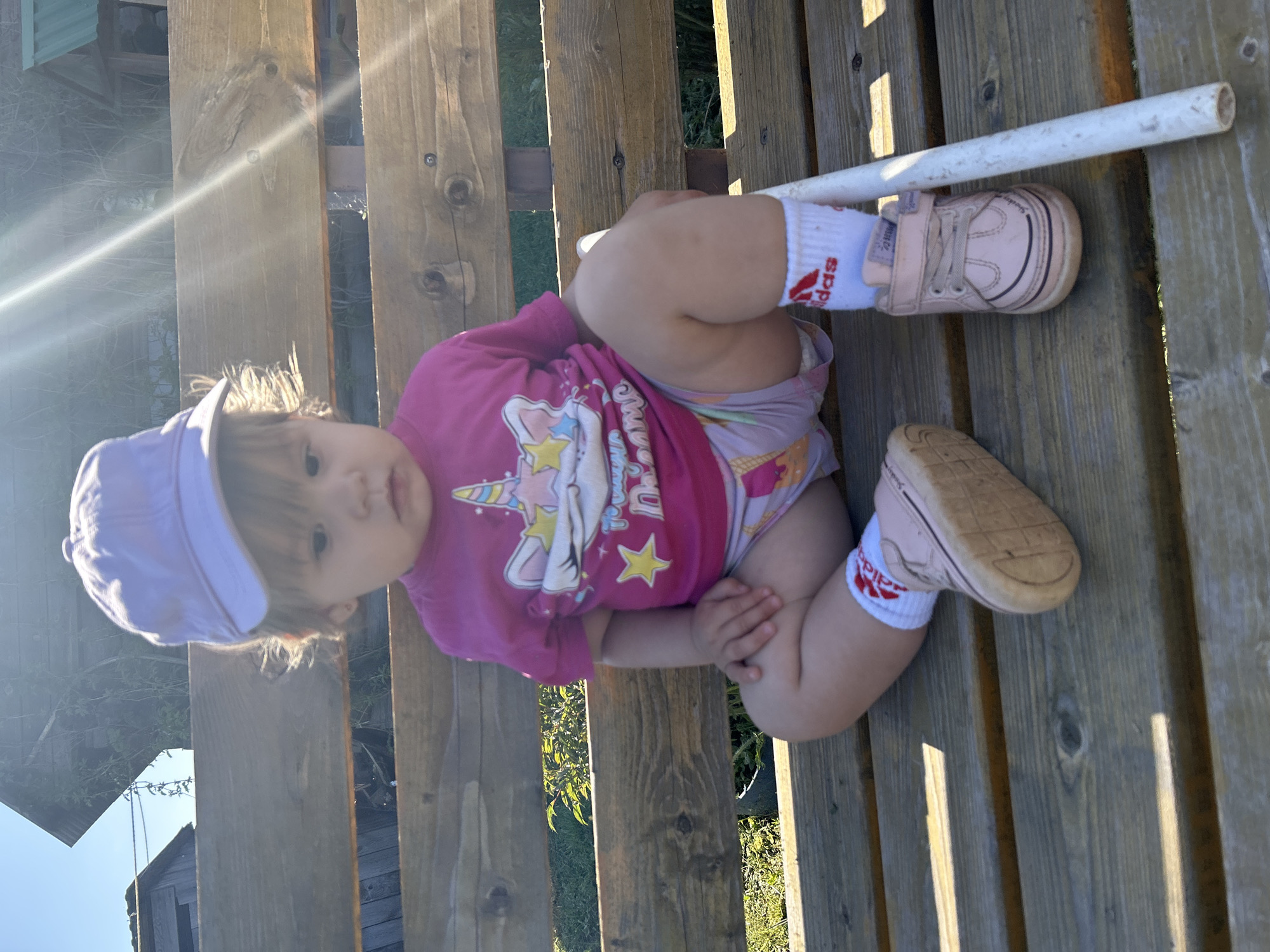 A toddler wearing a purple hat, a pink t-shirt with a unicorn design, light-colored underpants with a unicorn and star pattern, white socks with Adidas logos, and pink shoes is sitting on a slatted wooden surface, likely a bench. A white tube is positioned next to the toddler's feet. The scene appears to be outdoors in a sunny location, potentially in a backyard or park. In the background, there is a glimpse of a wooden structure, possibly a shed or a house, along with some greenery. This image was taken in the city of Ольховка, Russia.