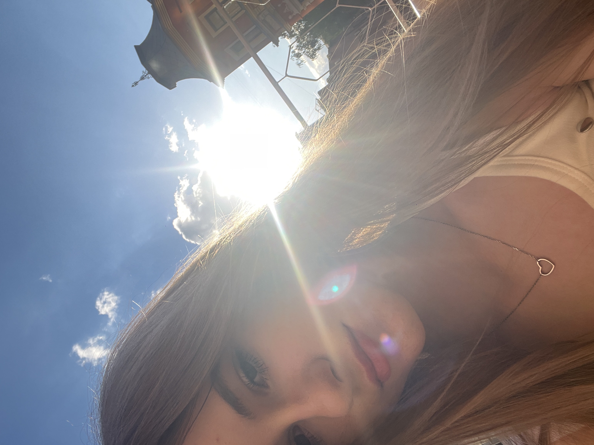 A selfie is taken outdoors on a sunny day with a bright blue sky and scattered clouds. The sun is high in the sky, creating lens flare effects across the image, obscuring some details. The perspective is from a low angle, looking up towards the sky and the subject.
The main subject is a young woman, whose face is partially visible. She has long, light brown hair that frames her face. Her eyes are dark and her lips are painted with a reddish-pink lipstick. She is wearing a light-colored top, possibly a tank top, and a delicate silver necklace with a heart-shaped pendant.
In the background, a building with reddish-brown architecture and visible windows can be seen against the sky. The architecture suggests an urban or town setting. The overall mood appears to be cheerful and relaxed, typical of a sunny day outdoors. The time of day is clearly midday, given the high position of the sun.
The image appears to be taken in Dnipro, Ukraine, based on the provided context. There is no visible text in the image.