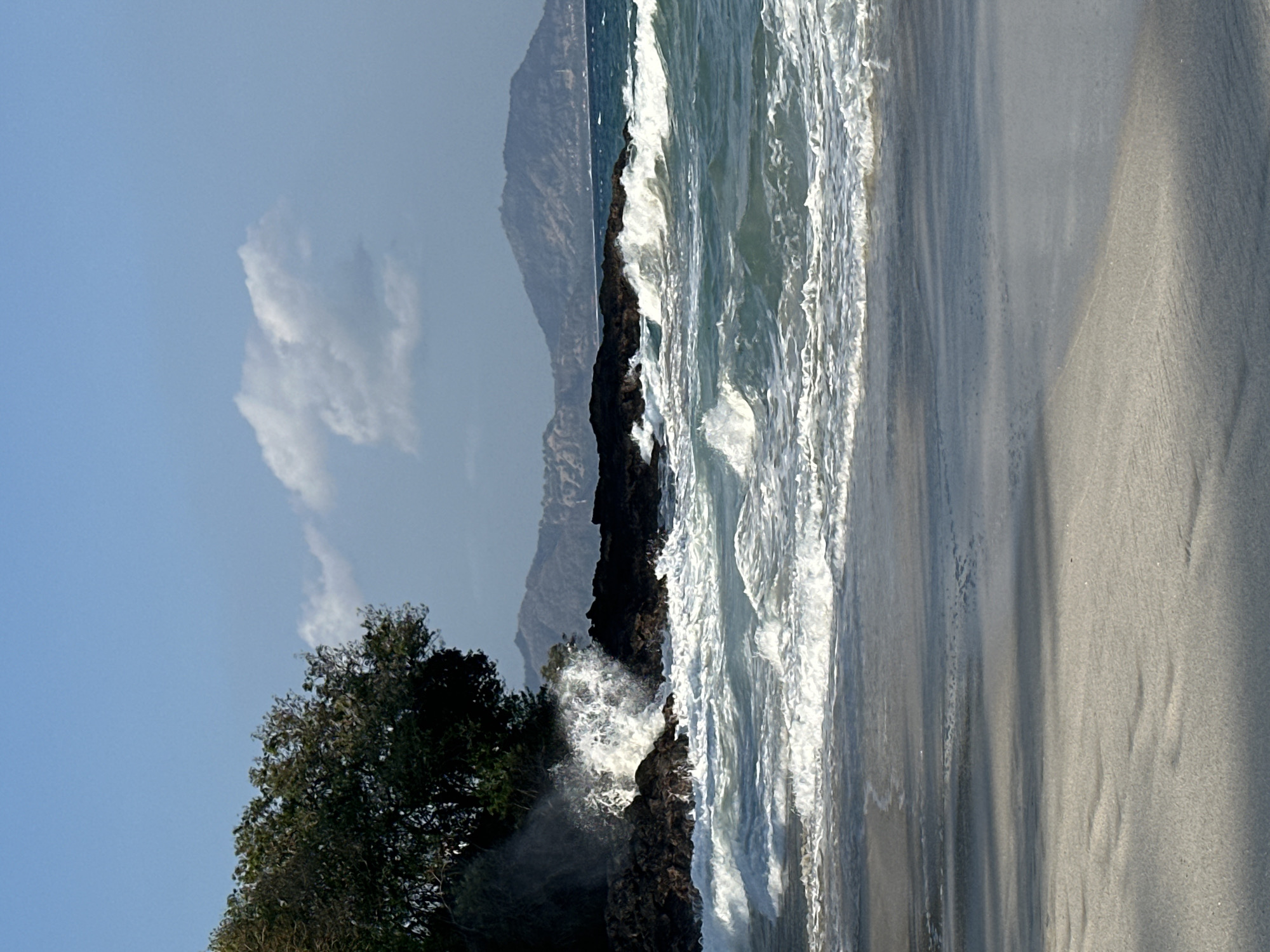 The image depicts a beach scene with waves crashing against dark rocks on the left side of the frame. A lush green tree is situated atop the rocks. The ocean is a vibrant turquoise, with whitecaps indicating active waves. In the background, hazy mountains are visible against a clear blue sky with a few scattered white clouds. The foreground consists of wet sand, reflecting the sky and creating a smooth, dark surface. The overall scene is natural and coastal, with no people or man-made structures immediately apparent in the immediate vicinity of the shore, suggesting it could be a secluded or natural beach area within Manggis, Indonesia, possibly near a volcanic coastline given the dark rocks.