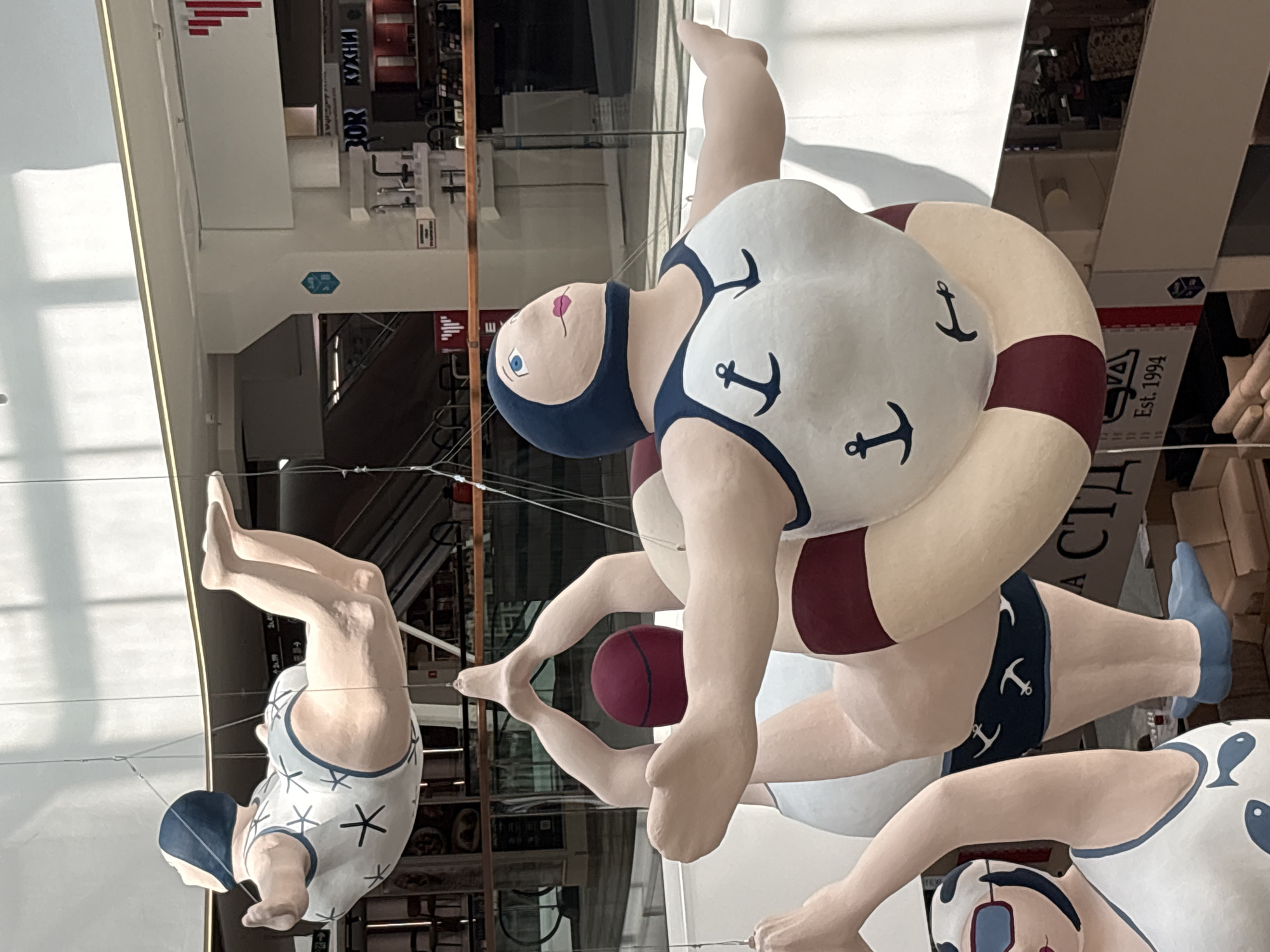 The media file showcases a whimsical art installation featuring oversized, stylized figures of swimmers suspended from above. The primary subjects are three larger-than-life figures, seemingly made of fiberglass or a similar material, dressed in vintage-style swimsuits. The figures are posed in dynamic, acrobatic positions reminiscent of synchronized swimming or diving.
One prominent figure, in the center, wears a white swimsuit adorned with dark blue anchors and a dark blue cap. A large, cream-colored life preserver with dark red bands encircles this figure. Another figure, to the left and slightly lower, wears a white swimsuit with a pattern of white starbursts and a dark blue cap, its legs bent at the knees as if mid-dive. A third figure, mostly visible at the bottom right, also wears a white swimsuit with blue trim and appears to be part of the ensemble. A dark red ball is visible between the legs of the central and left figures.
The background suggests an indoor setting, likely a public space such as a shopping mall or exhibition hall in Saint Petersburg, Russia. This is inferred from the architectural elements, lighting, and some visible signage. Text visible in the background includes "КУХНИ" (Kitchens) and "Est. 1994" on a box or sign, indicating a commercial or retail environment. The lighting suggests daytime, with bright overhead lights creating shadows and highlights on the figures and the surroundings. The overall mood is playful and surreal, with the unexpected scale and unusual poses of the swimmers creating a sense of wonder and amusement.