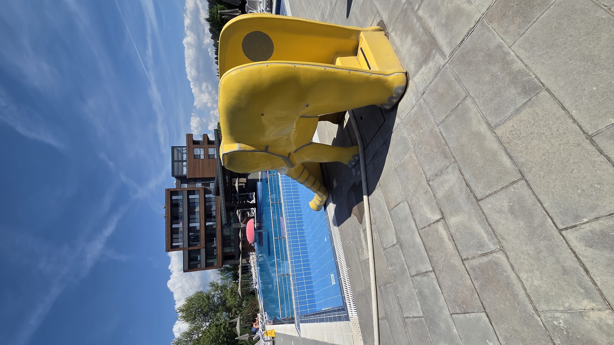 The image, rotated on its side, depicts a bright, sunny outdoor swimming pool area, characteristic of a resort or hotel in Bania, Bulgaria.
In the foreground and mid-ground, a large, bright yellow water slide, designed in the whimsical shape of a stylized animal (possibly a frog or cartoon character) with visible limbs, dominates the right side. The slide exhibits some wear and scuff marks. Adjacent to it is a clear blue swimming pool with a distinct tiled pattern on its bottom. The area surrounding the pool is paved with large, rectangular grey pavers. Indistinct figures are visible around the pool, some appearing to be relaxing on sun loungers. A red inflatable object floats in the pool, and a white pipe lies on the paved ground near the slide.
In the background on the left, modern multi-story buildings with dark frames, large glass windows, and sections of natural wood cladding suggest a contemporary architectural style. Green trees and foliage are also visible further in the background.
The sky is a vibrant blue with scattered wispy white clouds and faint contrails, indicating a bright and clear day, likely midday or early afternoon. The overall atmosphere is leisurely and indicative of a vacation setting. No discernible text is visible on any objects within the image.