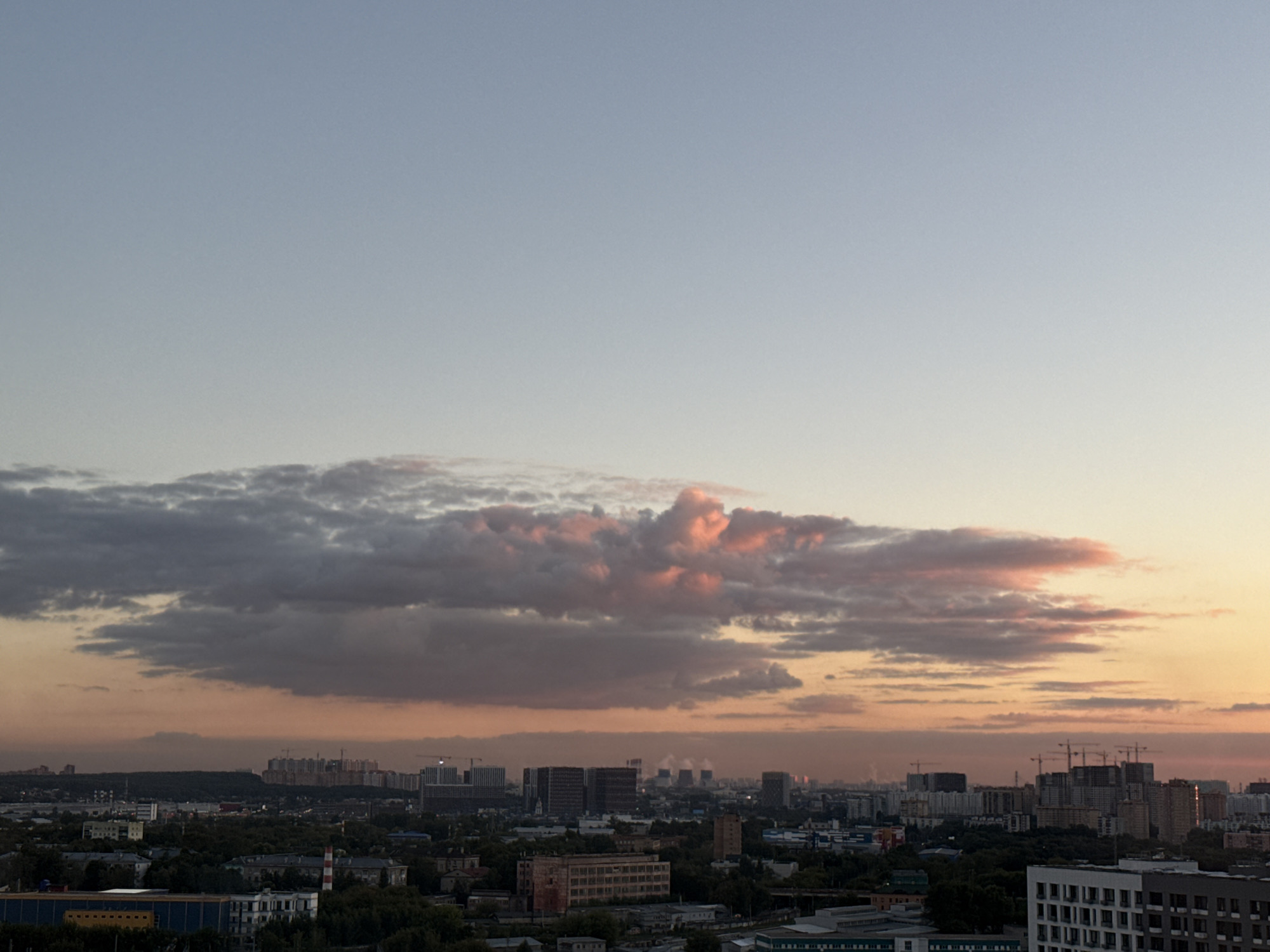 The image shows a panoramic view of Lyubertsy, Russia, at dusk. The sky is a soft gradient of blue and orange, with dramatic clouds in the upper left quadrant tinged with pink. The cityscape below is a mix of residential buildings, industrial structures, and construction sites, characterized by cranes and unfinished buildings. The foreground features a collection of modern apartment complexes, while the middle ground reveals a dense canopy of trees interspersed with older, brick buildings. In the distance, a vast urban sprawl stretches towards the horizon, with some buildings exhibiting illuminated windows. The overall atmosphere is one of a developing urban environment transitioning into nighttime.