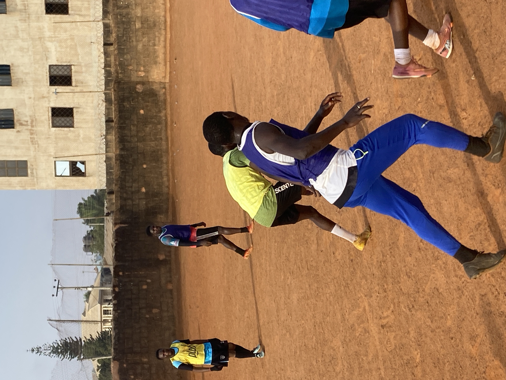 This image captures an outdoor scene in Barakin Lerawi, Nigeria, showing several young men engaged in athletic activity on a reddish-brown dirt field. The lighting suggests either early morning or late afternoon, with long shadows cast across the field, indicating a low sun angle and clear weather.
In the foreground and midground, multiple individuals are in various stages of movement, suggesting running, training, or playing a sport. A prominent figure in the center wears a purple and white tank top, blue track pants, and dark athletic shoes, captured mid-stride with arms bent. Behind him, another person in a neon yellow-green top with the partial text "SCENT E" and black shorts is also active. Further back, two other men are visible on the field; one wears a blue and black top with black shorts, and another in a yellow and blue top with the partial text "gold" and "DUO" also wears black shorts. A fifth person is partially seen at the top right, wearing a blue and light blue top with black shorts and pink shoes.
The background features a low, dark-colored wall, possibly concrete, topped with fencing that appears to have barbed wire. Beyond this wall stands a multi-story, light-colored building with numerous windows, some secured with bars. Further in the distance, trees, utility poles, and other structures are visible against a bright, clear sky, alongside a wire mesh fence. The overall scene depicts a casual athletic gathering in a semi-urban setting.