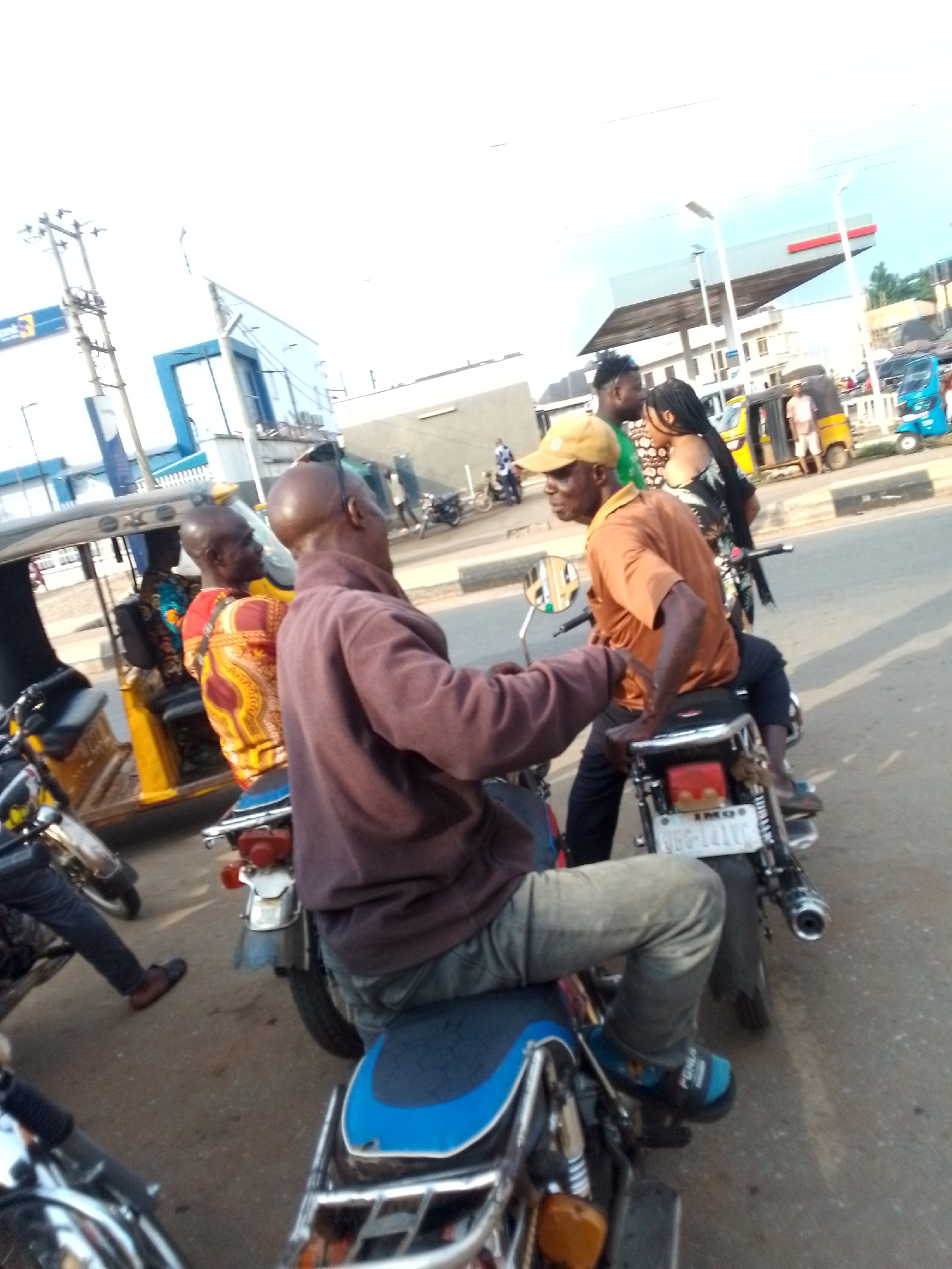 The image depicts a street scene in what appears to be Orlu, Nigeria. Several people are on motorcycles, stopped on the road. A man in a brown sweatshirt and light-colored jeans is facing another man on a motorcycle, who is wearing a yellow cap and an orange shirt. A license plate that reads "IMO URS-141VC" is visible on one of the motorcycles. In the background, there are buildings, an auto rickshaw (Keke Napep) and other motorcycles. A gas station is visible further in the background. The overall scene suggests a busy street or intersection.