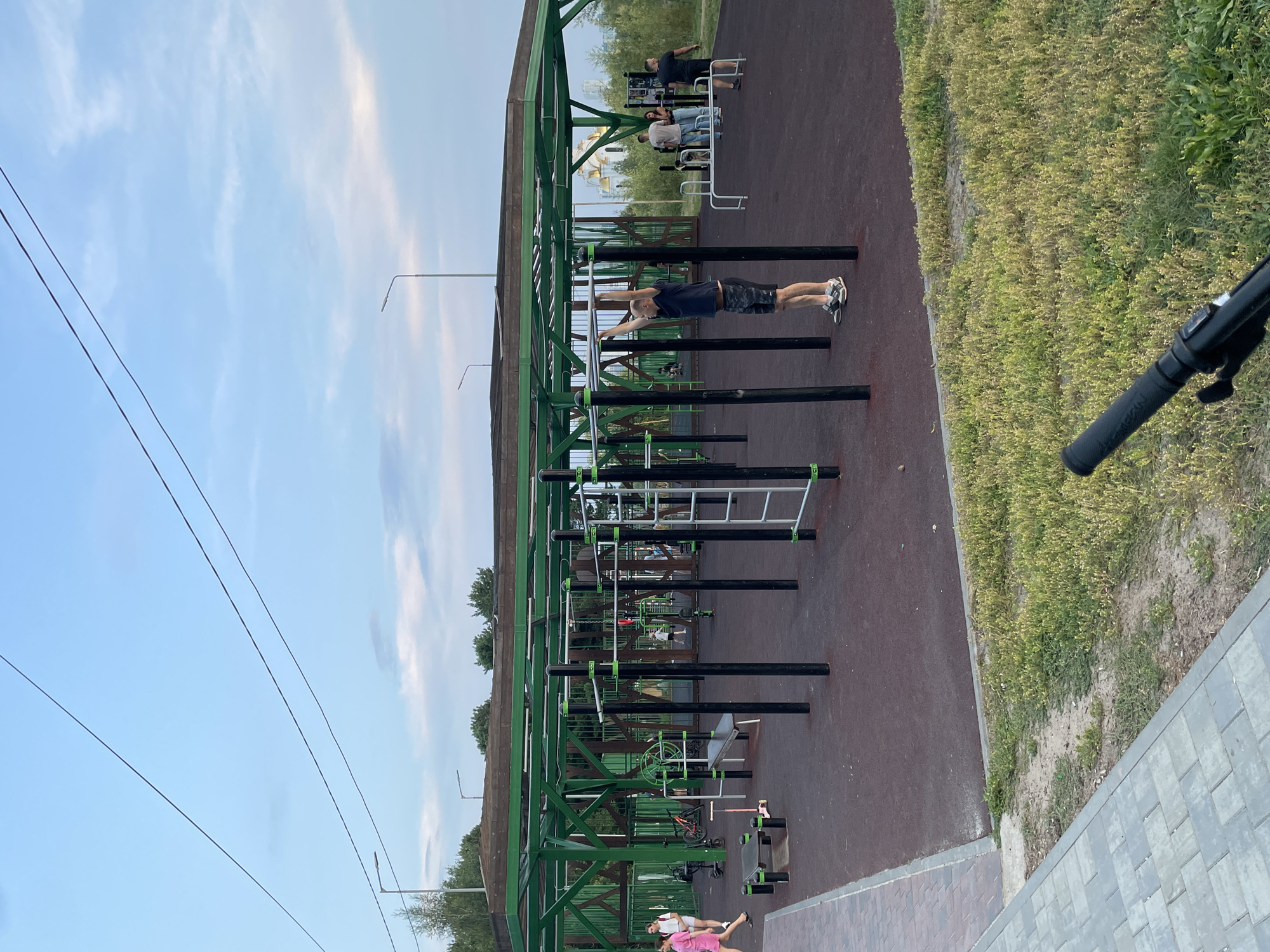 The image depicts an outdoor fitness park in Surgut, Russia, possibly located near the Church of the Savior Transfiguration based on the distinct golden domes visible in the background. The park features a covered area with numerous exercise stations, including parallel bars, pull-up bars, and what appears to be a leg press machine. Several individuals are present, with one man actively engaged in a pull-up exercise. Others are standing around or walking, suggesting a public recreational space. The ground surface is a dark reddish-brown, possibly rubberized for safety. The surrounding environment includes green trees and a cloudy sky. The overall scene suggests a well-maintained public facility for physical activity.