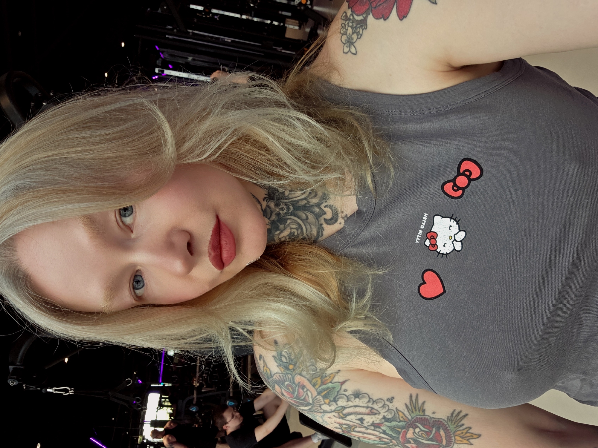 The image shows a fair-skinned person with blonde hair and blue eyes, wearing red lipstick. They have visible tattoos on their neck, arms, and upper chest. They are wearing a gray tank top adorned with Hello Kitty and heart graphics. The background appears to be a gym or fitness center, with workout equipment and other individuals visible in the blurred background. It is not possible to verify the location in the city of Saint Petersburg, Russia from the image.