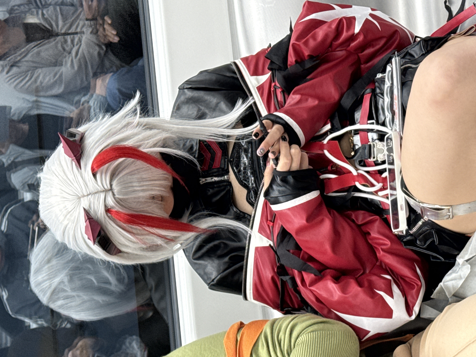 A person wearing a white wig with red streaks and a red and black jacket with white accents is the focal point of the image. The individual is also wearing black gloves with painted nails. The backdrop appears to be a reflection in a window or a glossy surface, showing indistinct shapes of people. The person is wearing green pants and there is a complex arrangement of straps, buckles, and other accessories attached to their clothing. The precise location cannot be determined as the surroundings are indistinct, but the prompt specified it is in 龙溪街道, China.