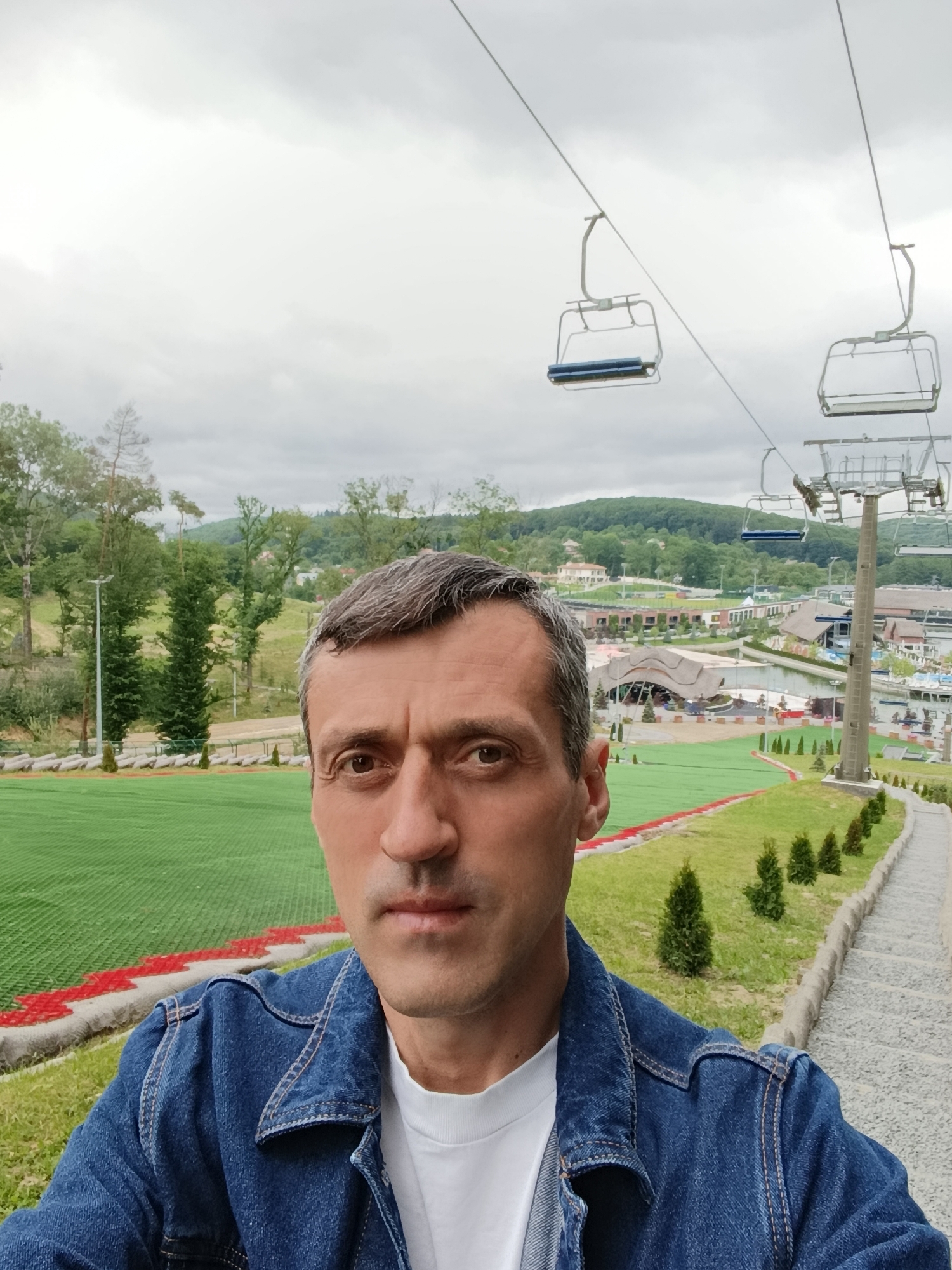 A selfie-style portrait captures a middle-aged male with short, dark hair exhibiting visible grey at the temples, looking directly at the camera with a neutral expression. He is wearing a blue denim jacket over a white crew-neck t-shirt. The background depicts an outdoor recreational area in Vynnyky, Ukraine, under an overcast sky. To the man's left, a wide expanse of bright green artificial turf, marked with red and white borders, extends, resembling a dry ski slope or summer tubing track. Beyond this, a treeline and a light pole are visible. To the right, a stone pathway lined with small, conical evergreen shrubs ascends. Overhead and to the right, multiple empty ski lift chairs are suspended from cables, supported by a prominent metal ski lift tower. In the mid-distance, a complex of buildings, some featuring thatched roofs, is visible adjacent to a body of water. Distant rolling, forested hills complete the horizon.