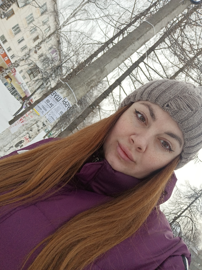 An outdoor selfie captures a woman in a winter urban setting in Chaikovsky, Russia. The woman, positioned prominently in the foreground, has long reddish-brown hair, wears a grey knit beanie, and a purple puffer jacket. Her face, seen from a slight upward angle, displays visible freckles, winged eyeliner, and a neutral expression with slightly parted lips.
The background is an overcast winter scene. To the left, a light-colored, multi-story apartment building with numerous windows is visible, partially covered by snow on the ground level. A red sign, characteristic of a commercial establishment (likely Pyaterochka supermarket), is partially visible on the ground floor. To the right, a concrete utility pole stands, adorned with multiple paper advertisements written in Cyrillic, attached with string. One poster clearly displays "АПРЕЛЬ" (April pharmacy) and "ул. Ленина, 3" (Lenin St., 3), indicating the precise location of the building in the background as Ulitsa Lenina, 3, Chaikovsky. Bare deciduous trees, stripped of leaves, frame the pole and the building against the grey sky, with snow covering the visible ground.