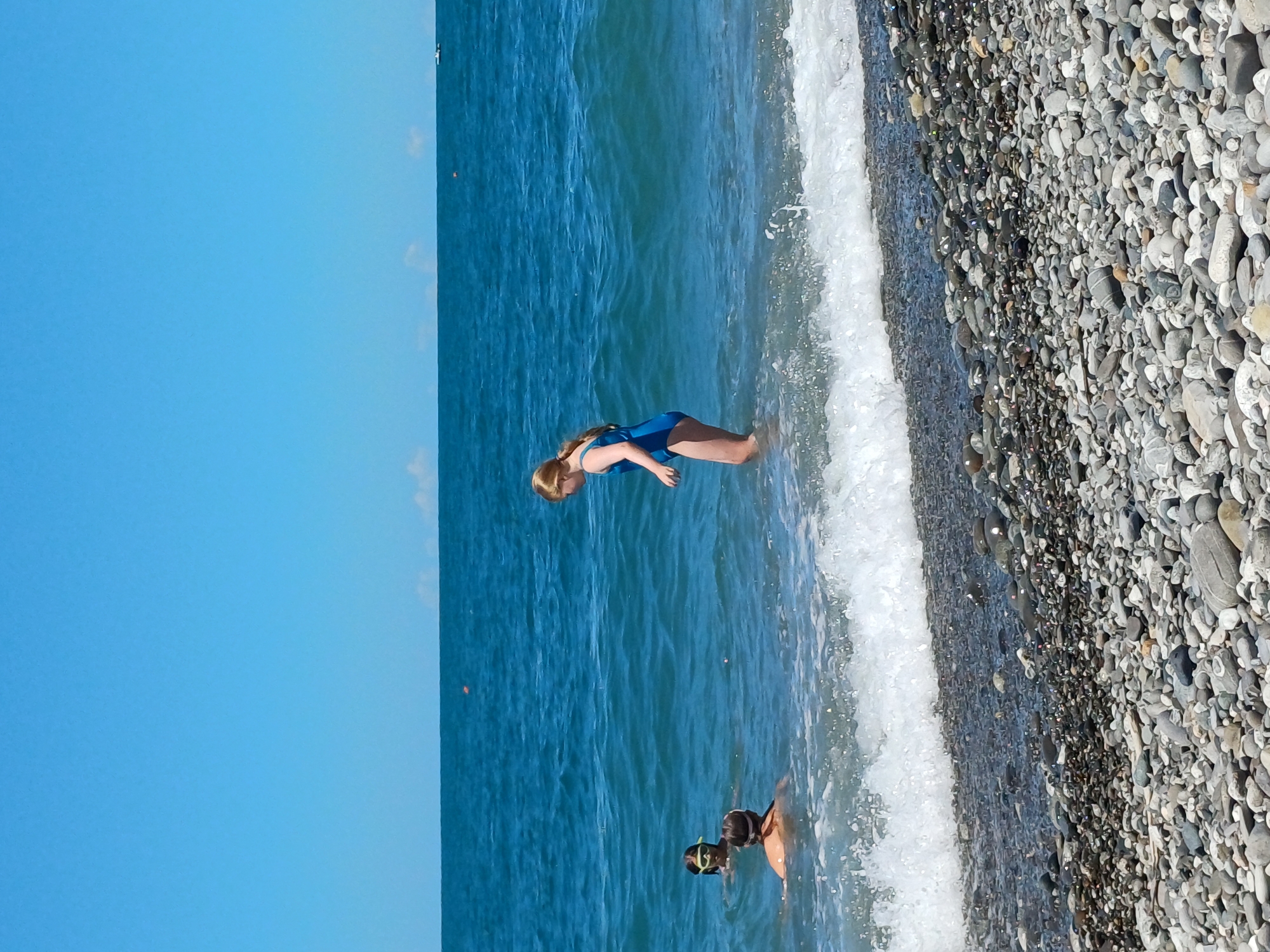 The image depicts a scene at a pebble beach in Sochi, Russia, under clear blue skies, suggesting a sunny day. The primary subjects are people engaged in beach activities. In the foreground, a person in a blue swimsuit is partially submerged in the shallow water, appearing to be in motion, possibly mid-dive or movement. Further back, two individuals are in the water, one wearing a snorkeling mask, indicating swimming or water exploration. The scene is divided by the shoreline, with the ocean occupying the left side and a dense covering of grey and white pebbles making up the right. Waves are breaking on the shore, creating white foam that contrasts with the deep blue of the water. The overall mood is one of leisure and outdoor recreation. No visible text is present in the image.