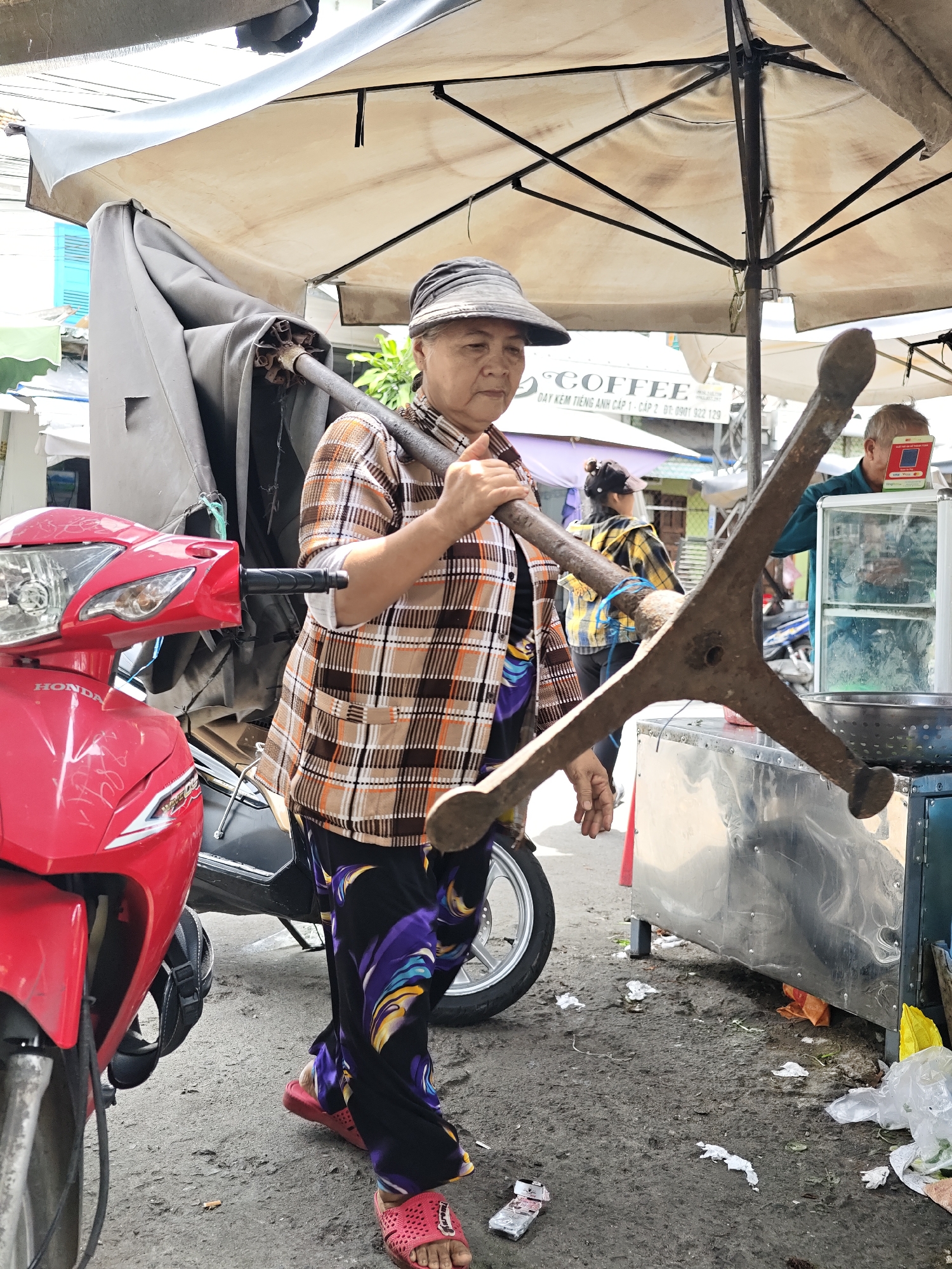 An elderly Asian woman, wearing a plaid shirt, colorful pants, a hat, and pink sandals, carries a long metal pole with a three-pronged attachment across her shoulders. She is walking on a dirt path lined with various stalls under awnings. A red Honda motorcycle is parked to her left. In the background, other people are visible, and a vendor is working behind a metal counter. The scene appears to be a market or street vendor area in Nha Trang Ward, Vietnam, characterized by its bustling activity and casual setup.