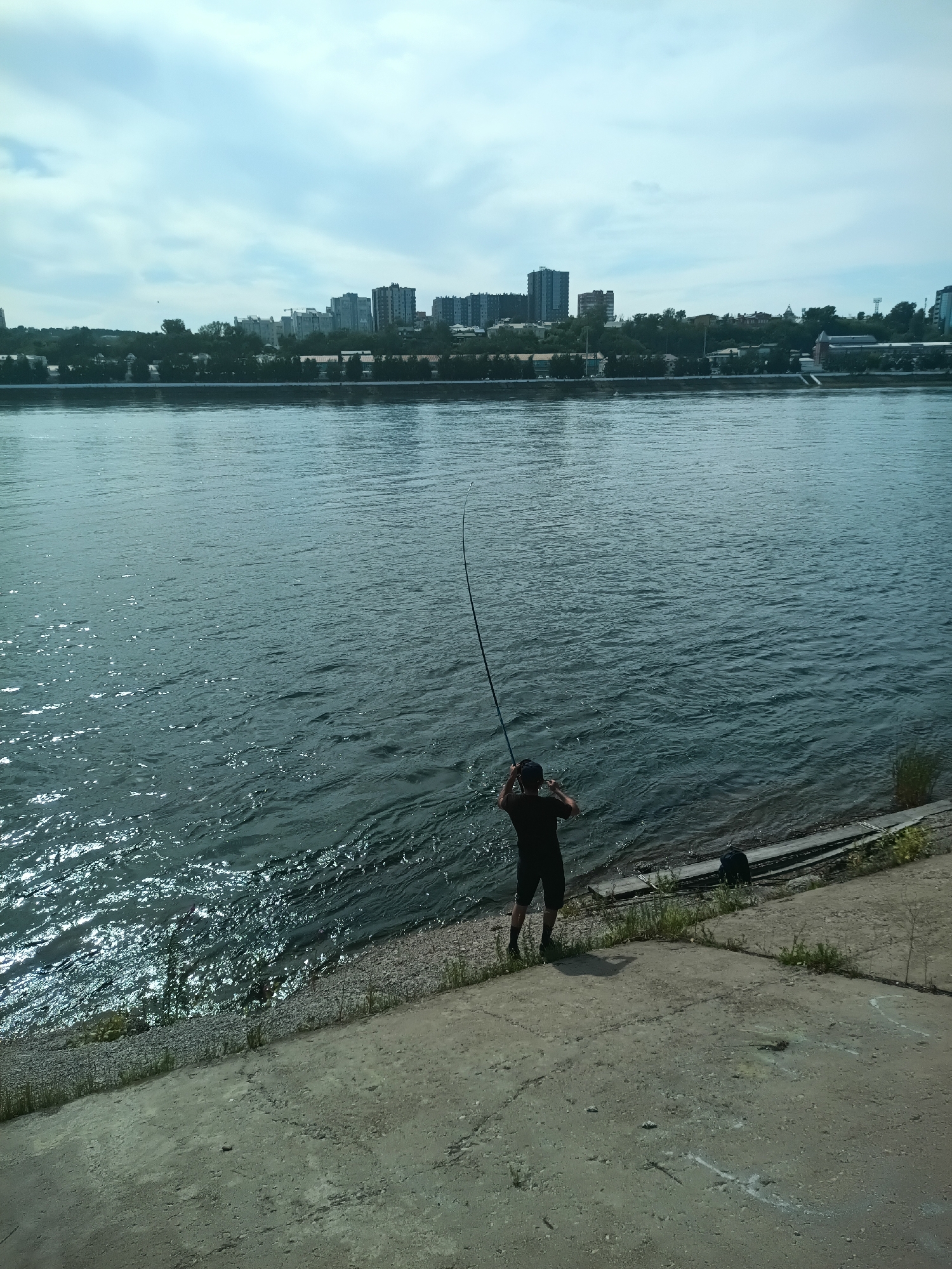 A lone individual is depicted fishing on the bank of the Angara River in Irkutsk, Russia, with a city skyline visible in the background. The person, dressed in a black t-shirt and shorts, stands facing the water, holding a fishing rod that curves significantly, suggesting it is bent from a catch or tension. The river's surface is choppy, reflecting the bright sky and clouds. The immediate foreground consists of a concrete embankment, transitioning to a pebbled shoreline where the person is standing. A black backpack rests on the ground near the water's edge. In the distance, a line of buildings, including several tall apartment blocks, forms the urban landscape of Irkutsk, situated on the opposite bank of the river. The sky is overcast with patches of blue.