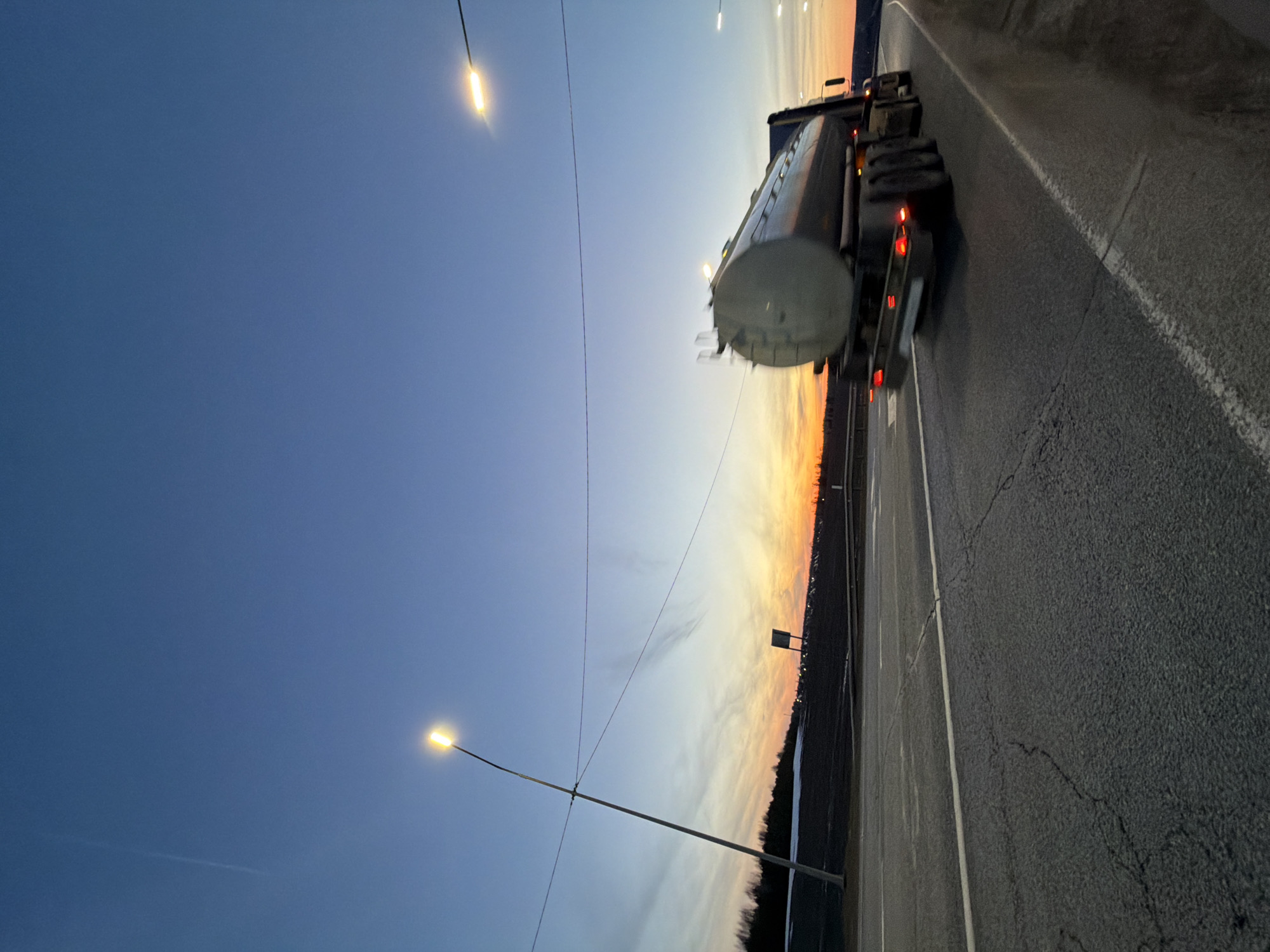 The image depicts a tanker truck driving on a paved road at dusk. The road appears to be a highway near Kuregovo, Russia, given the landscape and street lighting. A dark wooded area is visible alongside the road where the sun sets. The sky transitions from dark blue to orange near the horizon. Street lights illuminate the road, and the tanker truck's tail lights are visible. No other vehicles or people are evident in the frame.