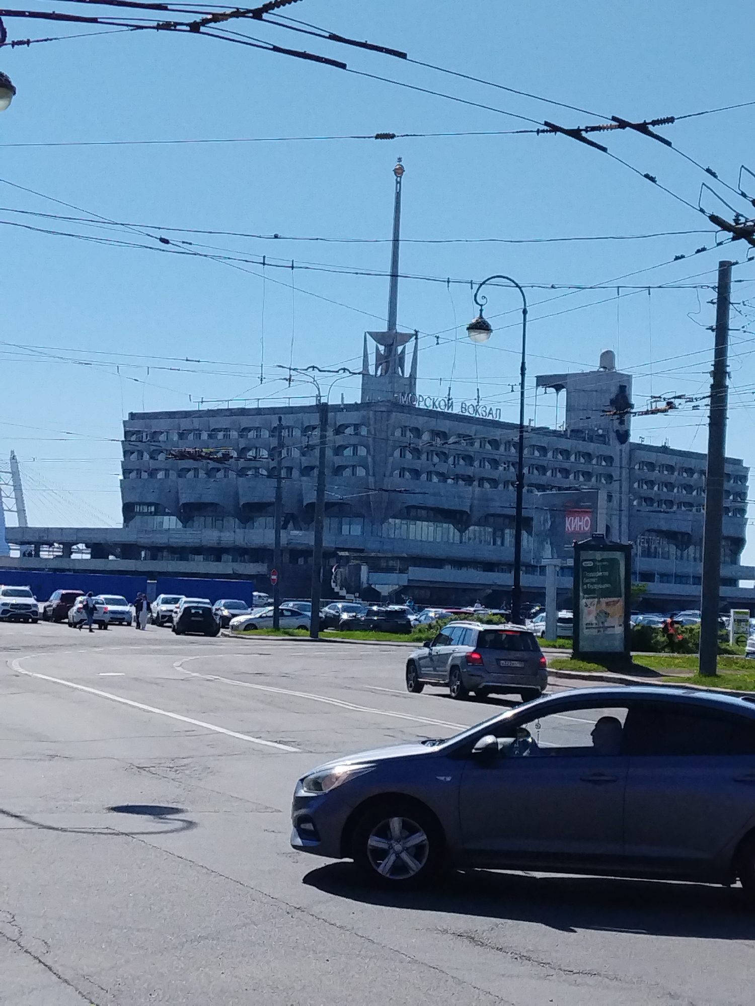 The image depicts a street scene in Saint Petersburg, Russia, featuring the Marine Facade passenger port (Морской вокзал). The building, a large, Brutalist-style structure with a prominent spire, is visible in the background. The Cyrillic inscription "МОРСКОЙ ВОКЗАЛ" (Marine Terminal) can be seen on the building's facade. A sign with "КИНО" (Cinema) is partially visible on the right side of the background.
Several cars are present in the foreground and midground, including a blue sedan in the immediate foreground. Other cars are parked in front of the port building.
Overhead electrical wires crisscross the sky. A section of the cable-stayed bridge (likely part of the Western High-Speed Diameter highway) can be seen in the distance on the left side.
The weather appears to be sunny, with a clear blue sky.