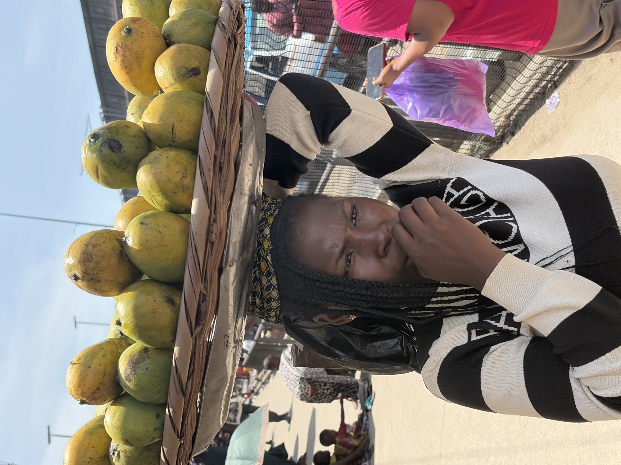 A young woman in Lekki, Nigeria, is carrying a woven basket of mangoes on her head. The basket is full of round, green and yellow mangoes. The woman is wearing a black and white striped sweater and has braided hair. She is looking at the camera and touching her face. In the background, there are other people and a metal fence. There is a person wearing a pink top purchasing the mangoes. There are other people selling goods. The lighting suggests it is daytime.