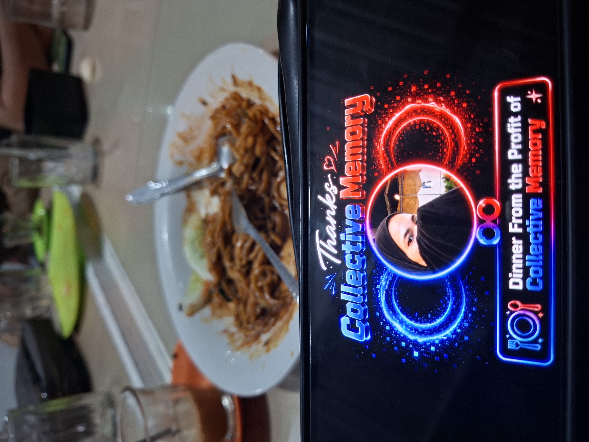 The scene shows a plate of stir-fried noodles on a table, with cutlery. A smartphone is held in front of the plate, displaying an image with text "Thanks Collective Memory" and "Dinner from the Profit of Collective Memory", along with a person's portrait in a circular frame and neon graphics. The location is implied to be Lhokseumawe, Indonesia, based on the provided context. The environment is a restaurant setting with glassware and other dining items visible on the table.