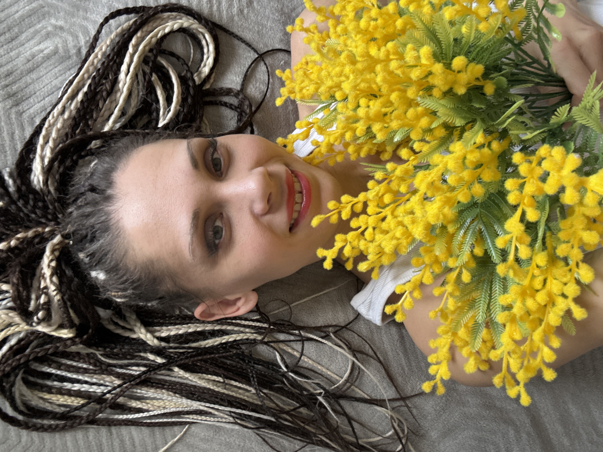 A woman lies on a gray surface in an unknown location in the city of Unknown, Unknown. She has brown and blonde braids. She is smiling and holding a large bouquet of yellow flowers. She is wearing a white top.