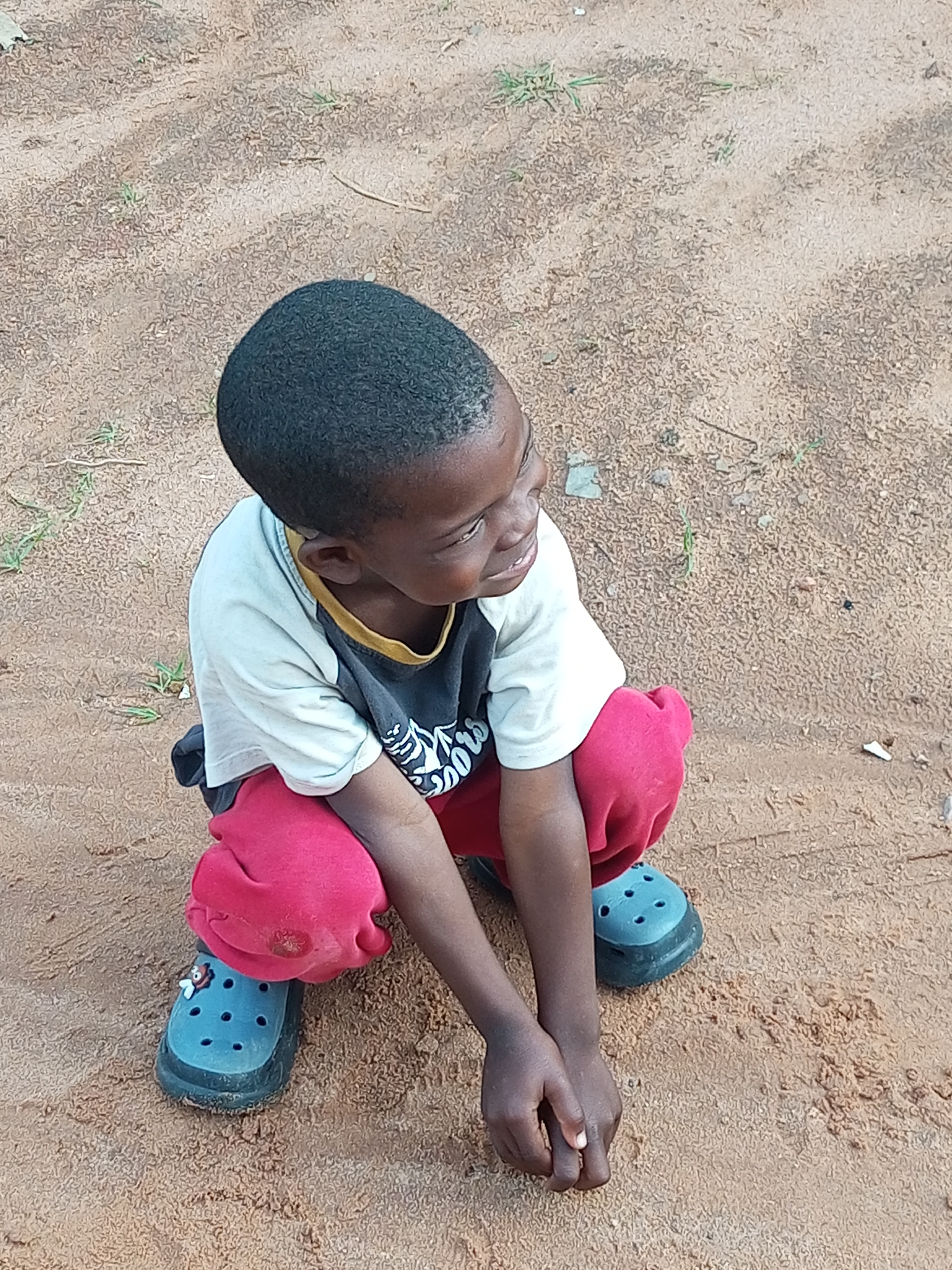 In a sandy outdoor environment, a young Black child, appearing to be around 4-6 years old, is crouched on the ground. The child has dark skin and short, dark hair. They are wearing a light-colored t-shirt with a black collar and a black graphic design on the front. Red pants are worn, and the child has blue Croc-style shoes on both feet, each adorned with a small, white and red charm. The child is looking to their right with their hands clasped in front of them. The ground is composed of light brown sand with sparse, dried grass and small pebbles scattered throughout. The image is an outdoor shot taken from a slightly elevated angle. The location is identified as Tempure, Nigeria.