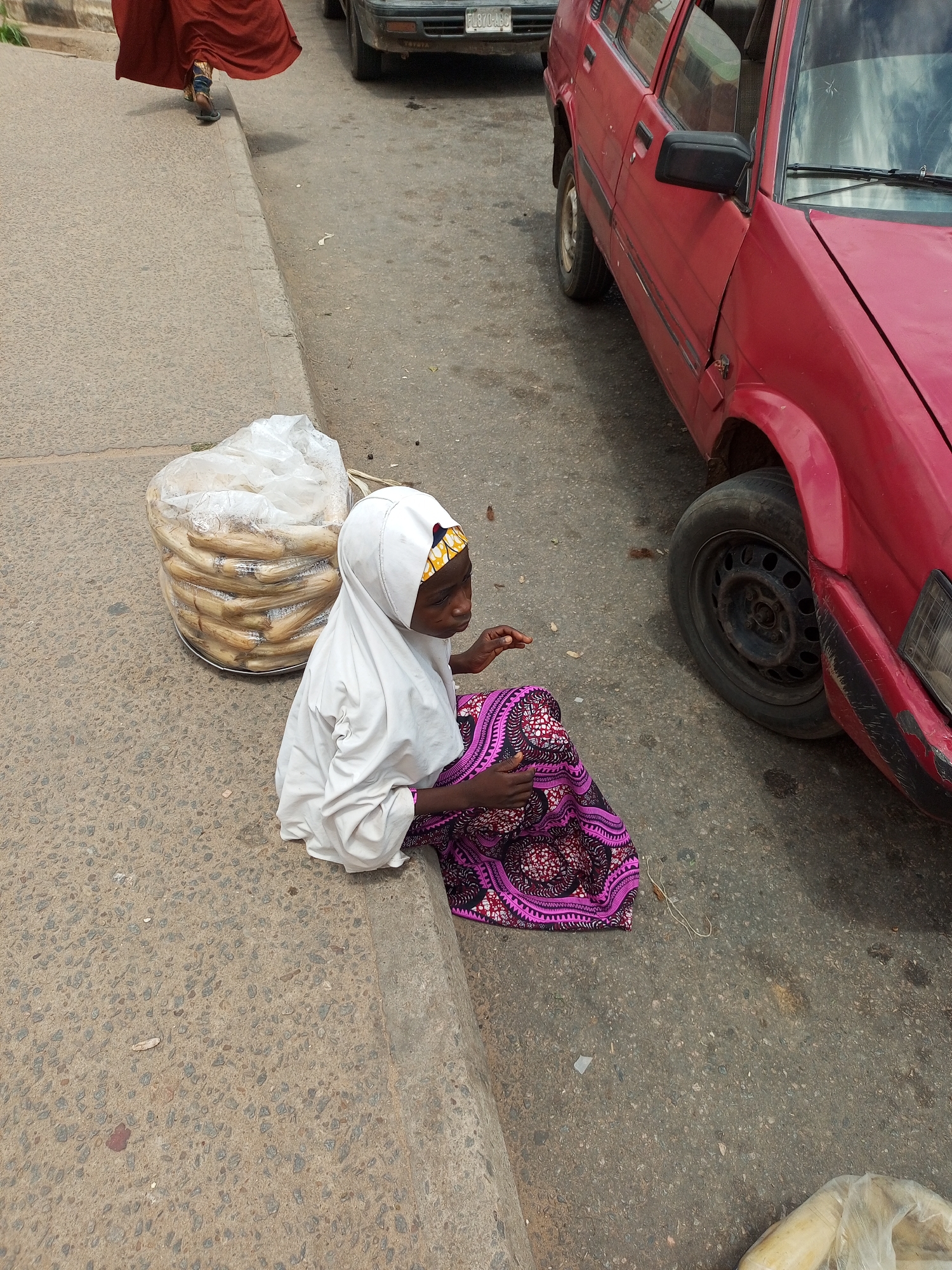 A young girl wearing a white hijab and a patterned pink and purple skirt sits on the curb of a street in Arawa, Nigeria. She is next to a large, clear plastic bag filled with what appears to be dried maize or corn. A red car is parked on the street behind her. In the background, a person in a red dress walks away from the camera. The street is paved with asphalt and there is a concrete curb. The overall scene depicts a typical street-side market or vendor setting.