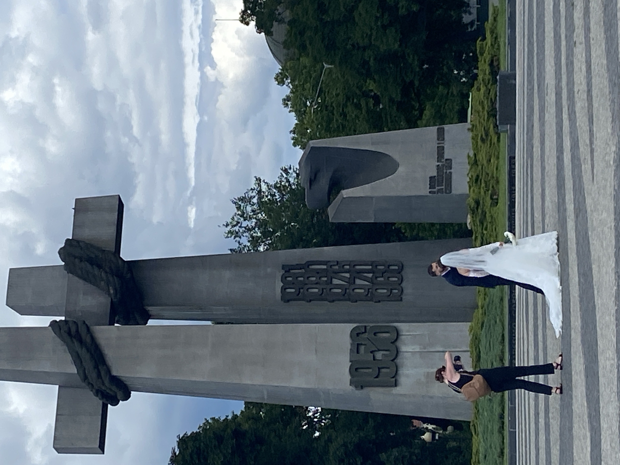 In Poznan, Poland, a wedding party is being photographed in front of a large, grey monument. The monument consists of two tall, angled pillars that form a cross shape, adorned with dark, rope-like sculptures. To the left of the cross, the year "1956" is inscribed. To the right, a column of dates including "1981", "1980", "1976", "1970", and "1968" are visible. A second, smaller monument with a bird-like head sculpture is visible behind the main monument.
In the foreground, a bride in a white wedding dress with a long train and a groom in a dark suit are standing close together, facing away from the camera. The bride is holding a bouquet. A woman with a backpack is standing to the left of the couple, holding a camera and appearing to take their photograph. The ground is paved with grey and white striped tiles. Lush green trees and a cloudy sky form the background.