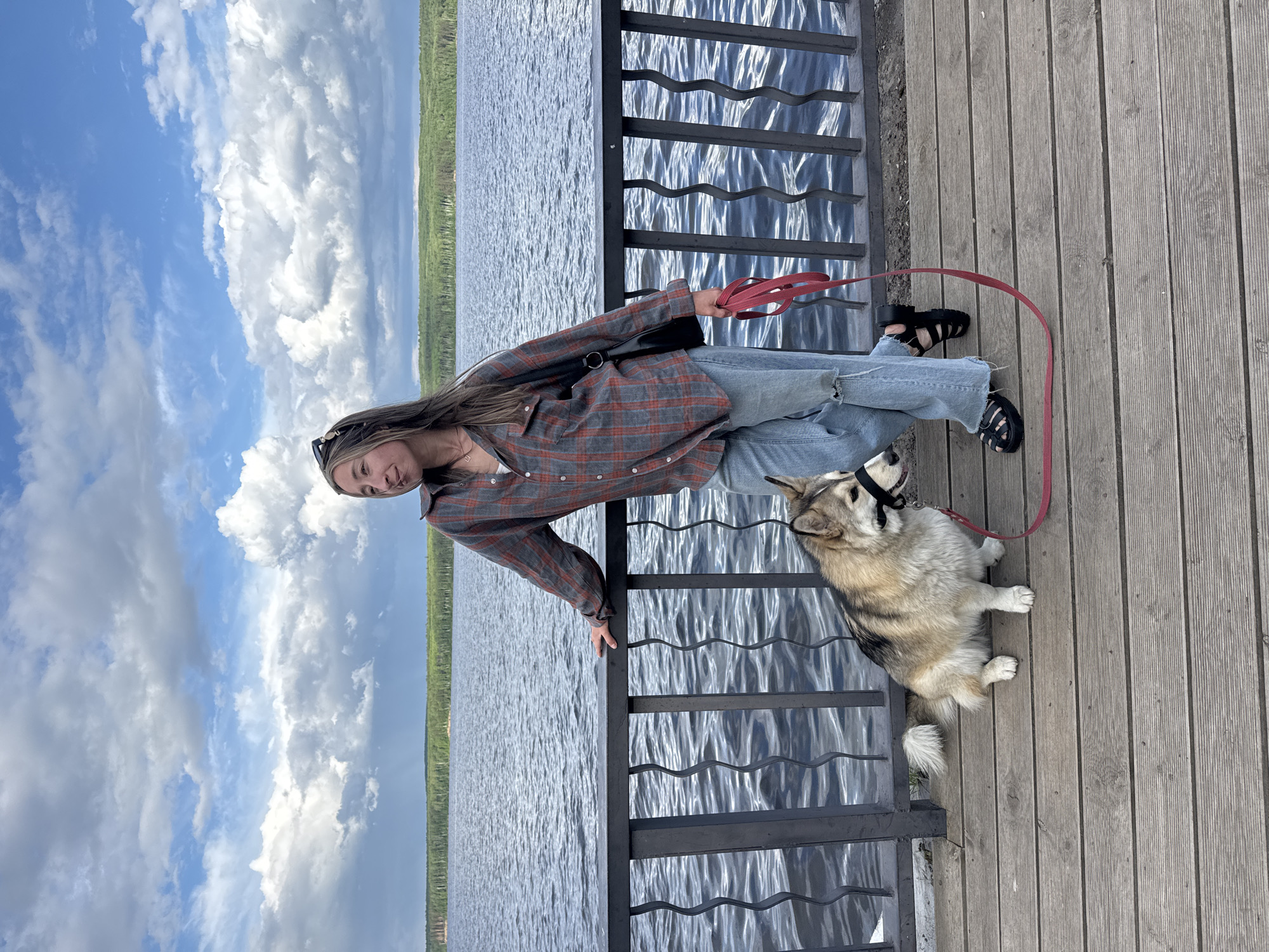 A young woman stands with a dog on a wooden pier, next to a metal railing, overlooking a body of water, likely in Chaikovsky, Russia. The woman is wearing a red and gray flannel shirt, ripped blue jeans, and black sandals. She holds a red leash. The dog, with a light brown and white coat and a black head halter, sits next to her. The background shows a lake, a strip of green trees on the far shore, and a blue sky with cumulus clouds. The scene is outdoors in daylight.