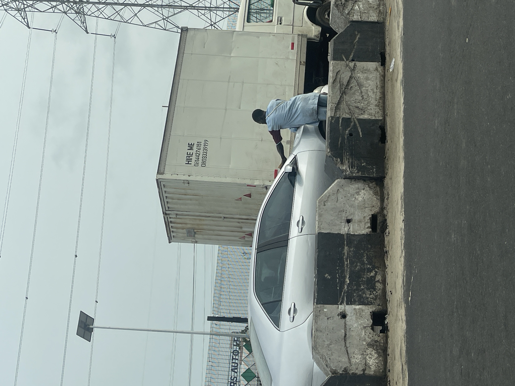 The image, captured in Igboefon, Nigeria, is presented with a rotated perspective, showing a daytime street scene under an overcast sky. In the foreground, a man, wearing a light blue tunic over dark trousers, is leaning into the open driver's side window of a silver or light-colored sedan, his back to the viewer.
Immediately behind the car and the man is a large white box truck or trailer. Printed prominently in black on its side are the words "HIRE ME" followed by two phone numbers: "08144276151" and "080333339199". To the right, concrete barriers, marked with alternating black and white sections, run alongside the asphalt road.
The background features numerous power lines stretching across the grey sky, supported by a large metal pylon structure visible in the upper left. A street sign or possibly a solar panel on a pole is also visible further down. Partially seen in the bottom left corner, on what appears to be a building or fence, are the letters "RS AUTO T". The scene suggests vehicles stopped or moving slowly in traffic on a busy road, possibly in an urban or industrial area.