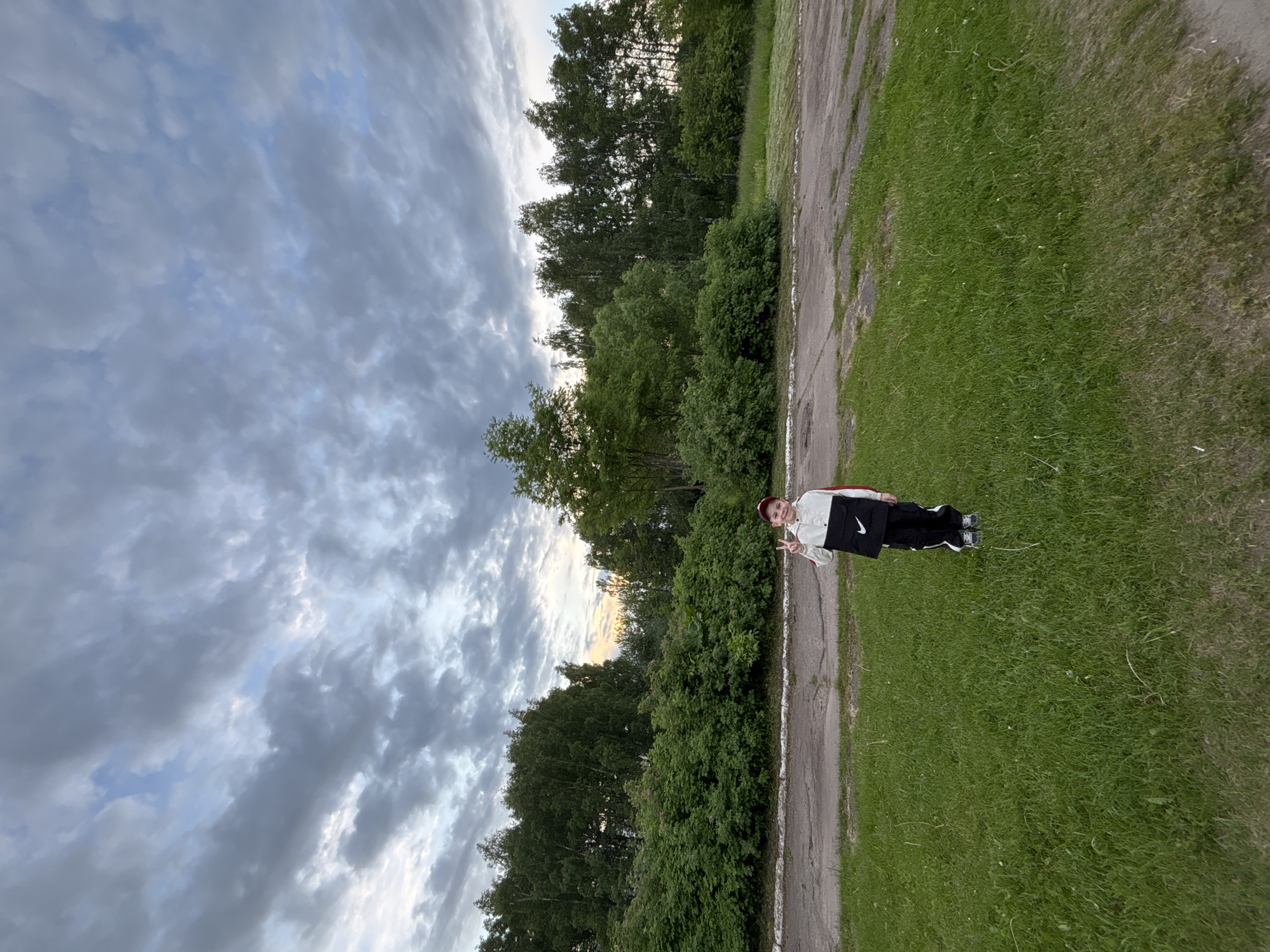 A full-body, outdoor shot depicts a young person standing on a worn dirt or asphalt path, making a peace sign with their right hand. The individual is positioned slightly off-center to the right, facing the camera. They are dressed in a two-toned jacket (cream on top, black on the bottom with a visible "Nike" logo), black pants with white vertical stripes, and dark shoes. A red beanie or hat completes their attire.
To the left of the path, a strip of green grass separates it from a dense line of trees and bushes that stretch across the background. The sky above is largely overcast with scattered, dark grey clouds, though patches of lighter sky with hints of orange and yellow light are visible on the horizon, suggesting late afternoon or early evening. The overall lighting is dim, consistent with cloudy weather. The scene appears to be in a natural or semi-natural environment, such as a park, forest edge, or rural area in Rusanovo, Russia. The person looks relaxed and happy.