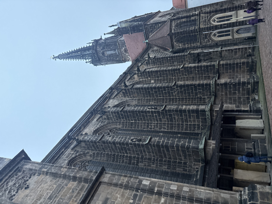 This image features a majestic, historic church or cathedral, viewed from a low angle looking upwards, likely depicting Meissen Cathedral in Germany. The primary subject is the colossal stone edifice, constructed from dark, weathered blocks, showcasing intricate Gothic architecture.
The main body of the church is characterized by robust buttresses and ornate window frames with delicate tracery patterns. Its side walls rise steeply, punctuated by rhythmic architectural elements. A reddish-brown roof section is visible where the main building transitions to a tower.
Dominating the background against a light, overcast sky is a very tall, slender spire. This spire is richly detailed with multiple tiers of pointed pinnacles and complex ornamentation, culminating in a cross at its apex. Along the roofline of the church's main structure, several dark, stylized figures, possibly gargoyles or decorative elements, add to its Gothic character.
The sky is a uniform, pale grey-blue, indicating an overcast or cloudy daytime setting without direct sunlight. In the lower right corner, several small figures are visible. One person, wearing a blue jacket, stands near large, light-colored stone blocks at the base of the church. Other figures are further in the background on what appears to be a paved area. Their attire suggests cool weather. No discernible text is visible within the image.
