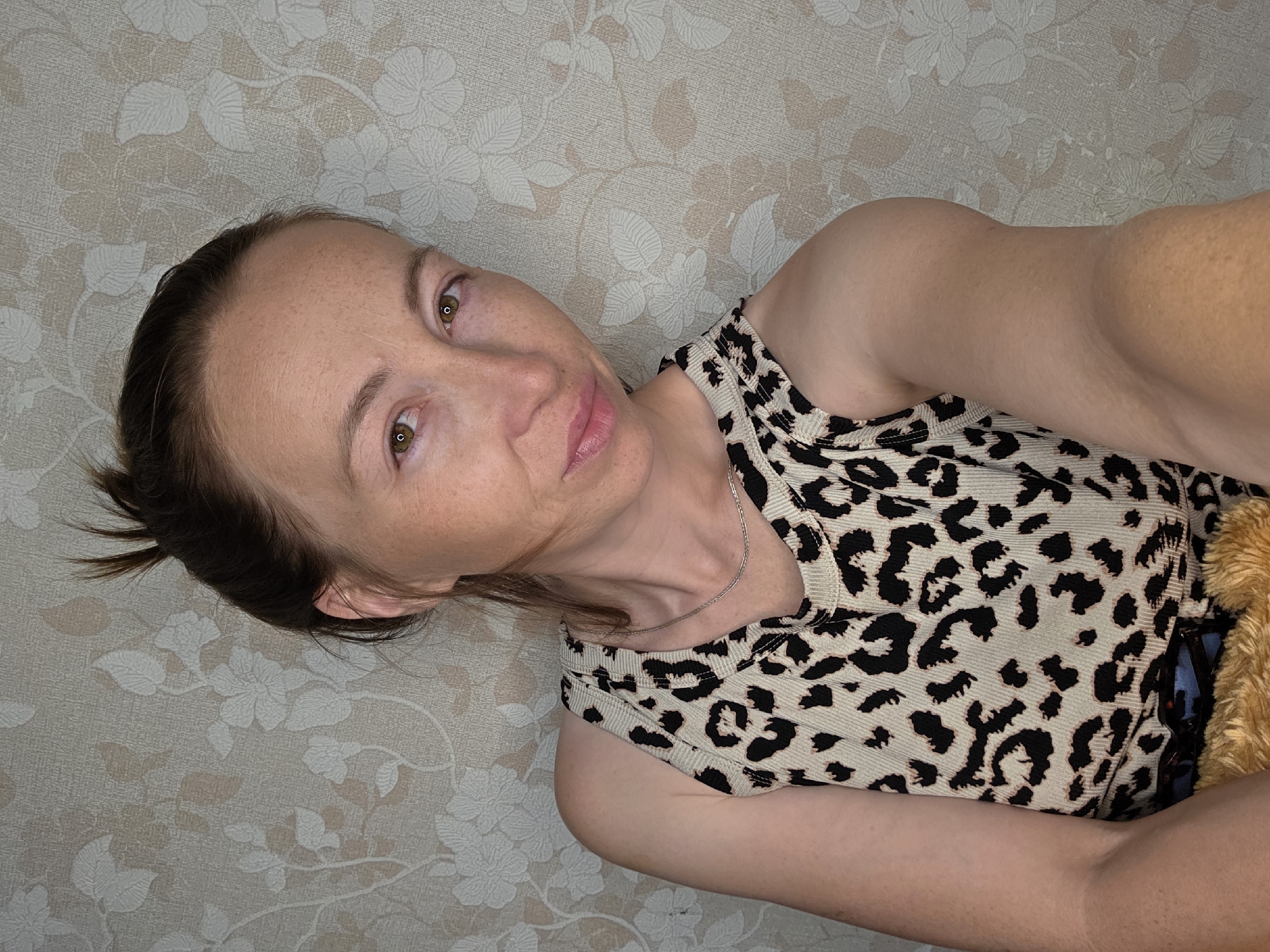 A medium shot, head and shoulders portrait of a young woman with light brown hair pulled into a messy bun is captured against a floral patterned wallpaper. She is looking directly at the camera with a neutral expression, her head tilted slightly to her right. The woman is wearing a sleeveless leopard print top and a delicate silver necklace. Her skin has a fair complexion with scattered freckles. Her eyes are a warm brown hue. The lighting appears to be from the front, casting a soft glow on her face. The background wallpaper is a light beige with a repeating pattern of stylized white and light brown flowers and leaves. The image is taken indoors, suggesting a home environment.
