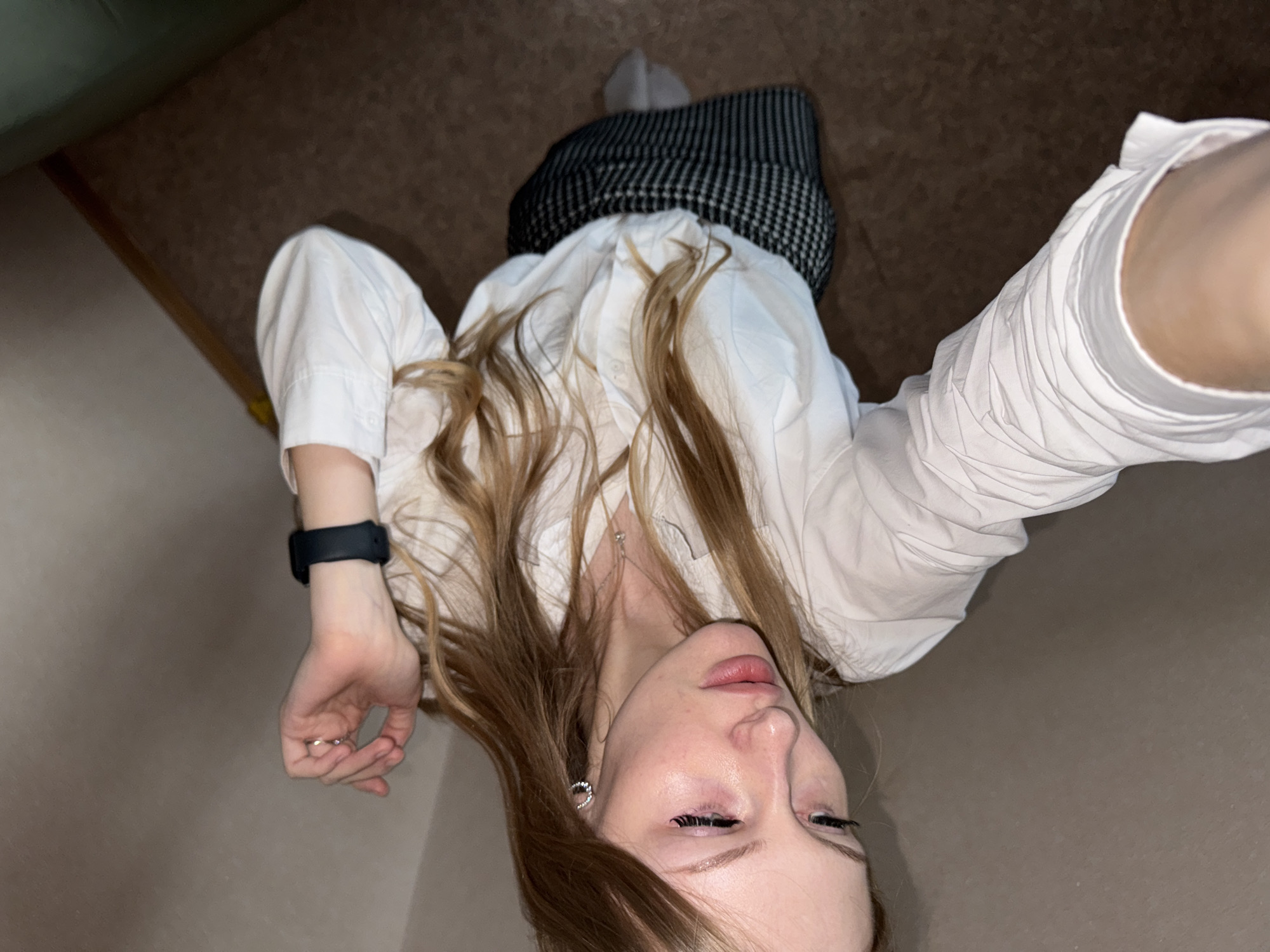 The image shows a young woman with blonde hair lying on her side on a carpeted floor. She is wearing a white button-down shirt and a black and white checkered skirt. A smartwatch is visible on her left wrist. The background appears to be an indoor setting, possibly a room in an apartment in the city of Perm, Russia, with a plain wall and a carpeted floor. The lighting suggests it is indoors.